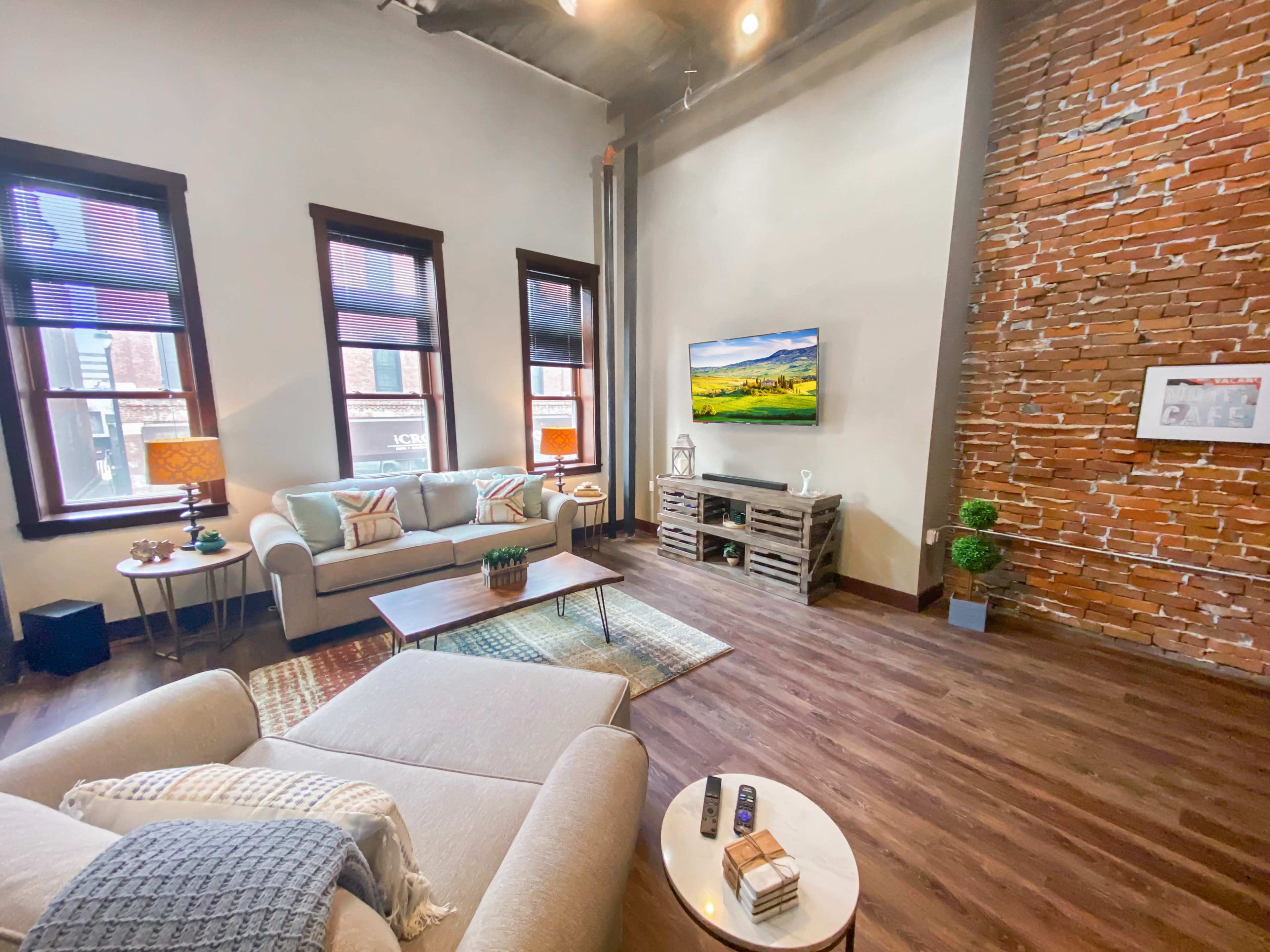 A modern living room featuring a beige sofa, a wooden coffee table, a TV mounted on a brick wall, and large windows with natural light.