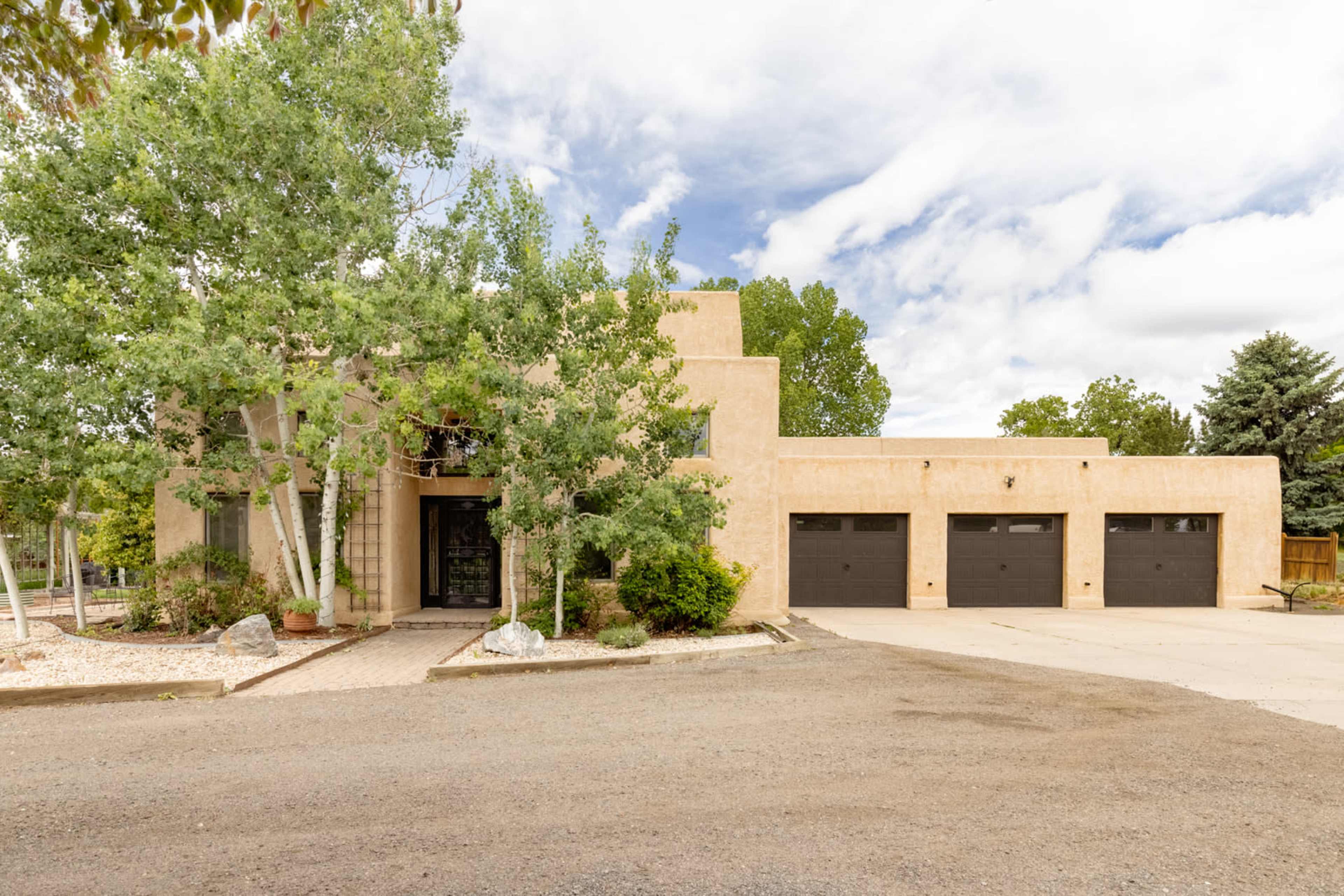 The image shows a single-story, adobe-style house with a front driveway and three garage doors, surrounded by trees and shrubs.