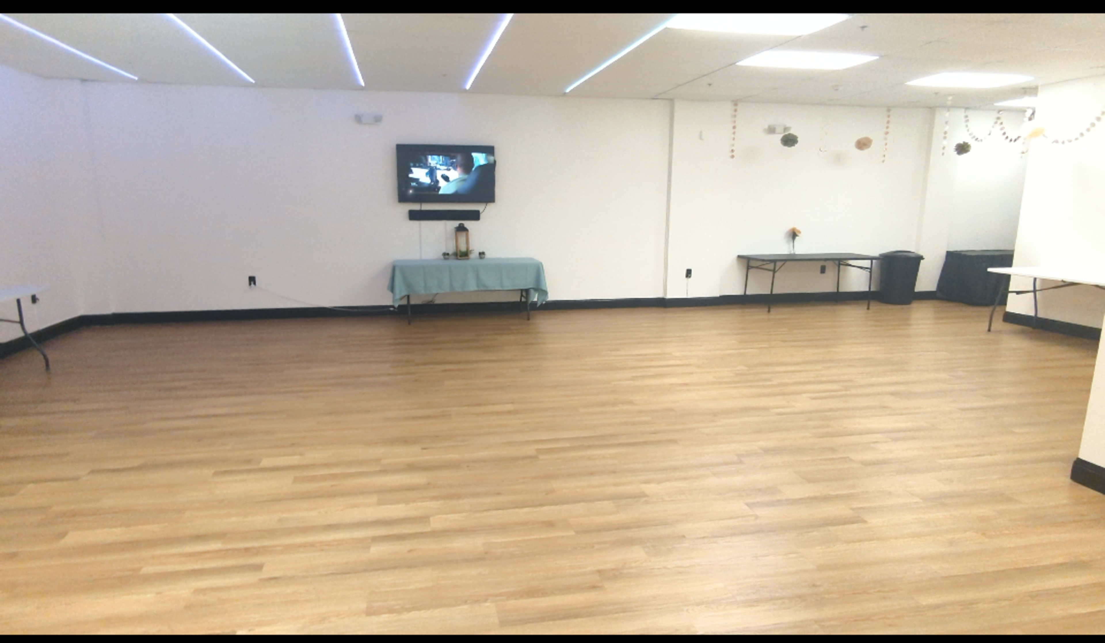 The image shows an empty room with wooden floors, a wall-mounted television, and several tables along the sides.