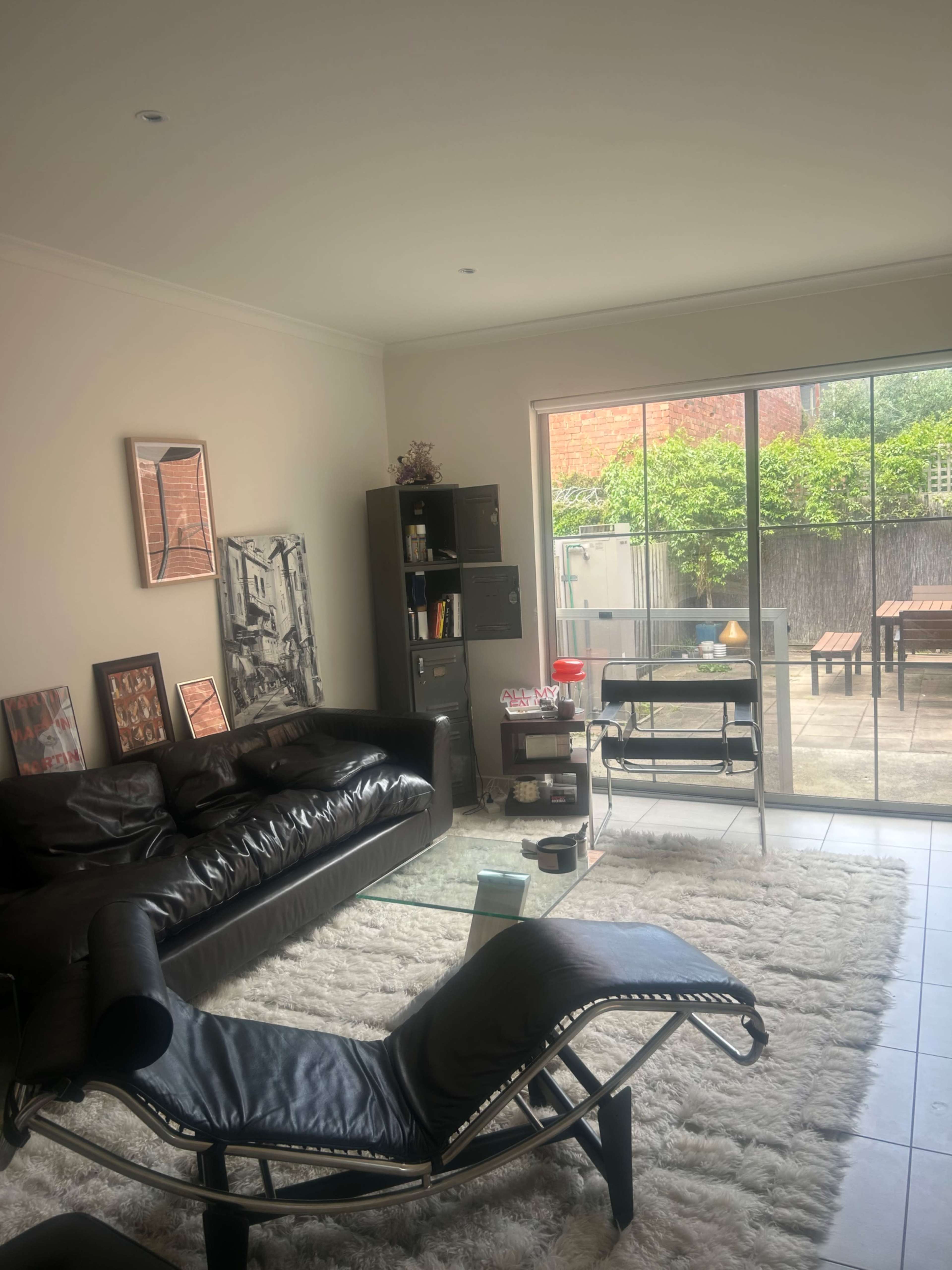 The image shows a modern living room with a black sofa, a glass coffee table, a lounge chair, and large windows facing an outdoor patio area.