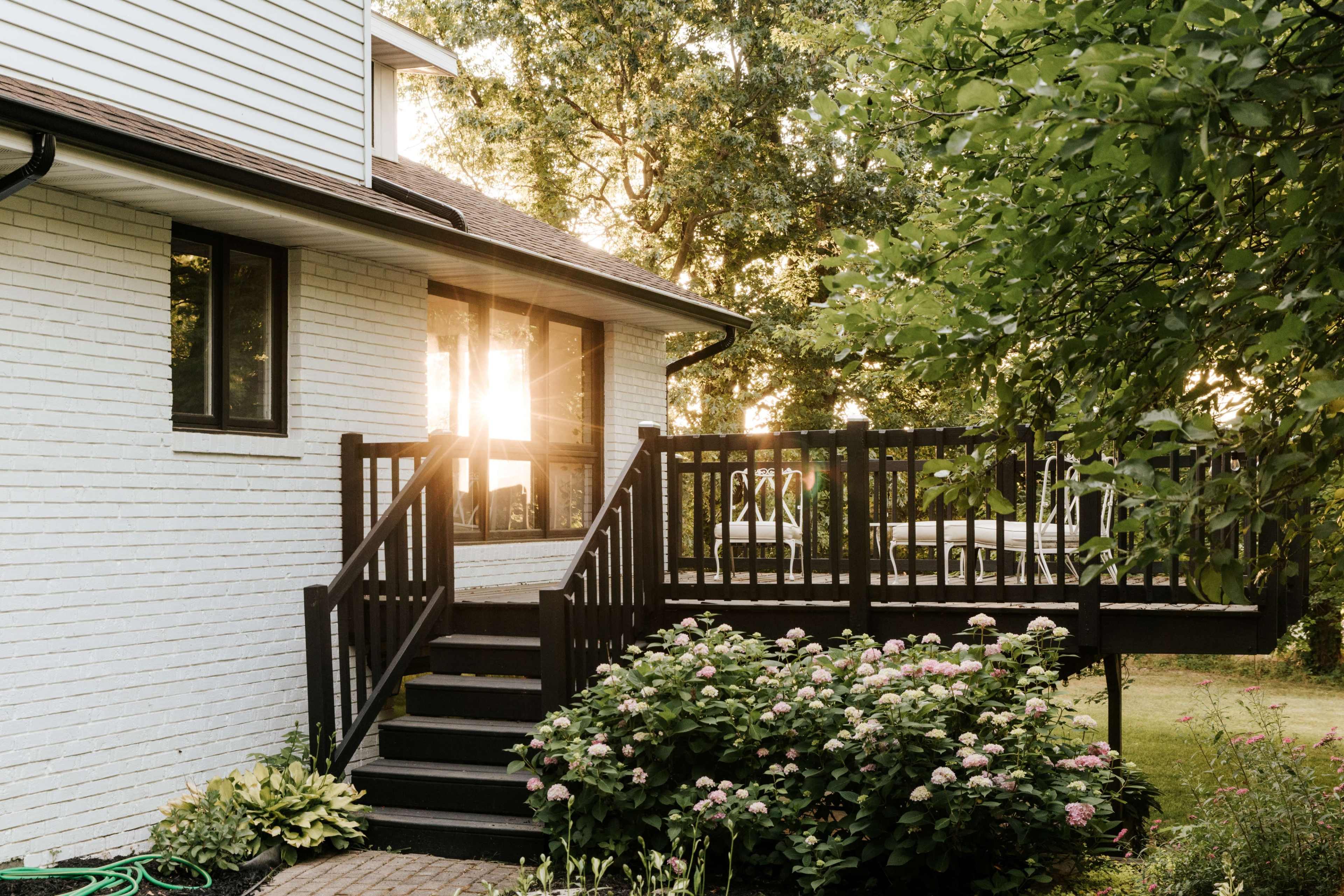 The image shows a wooden deck with stairs leading up to a house, surrounded by green foliage and flowers in the foreground, with sunlight filtering through the trees.