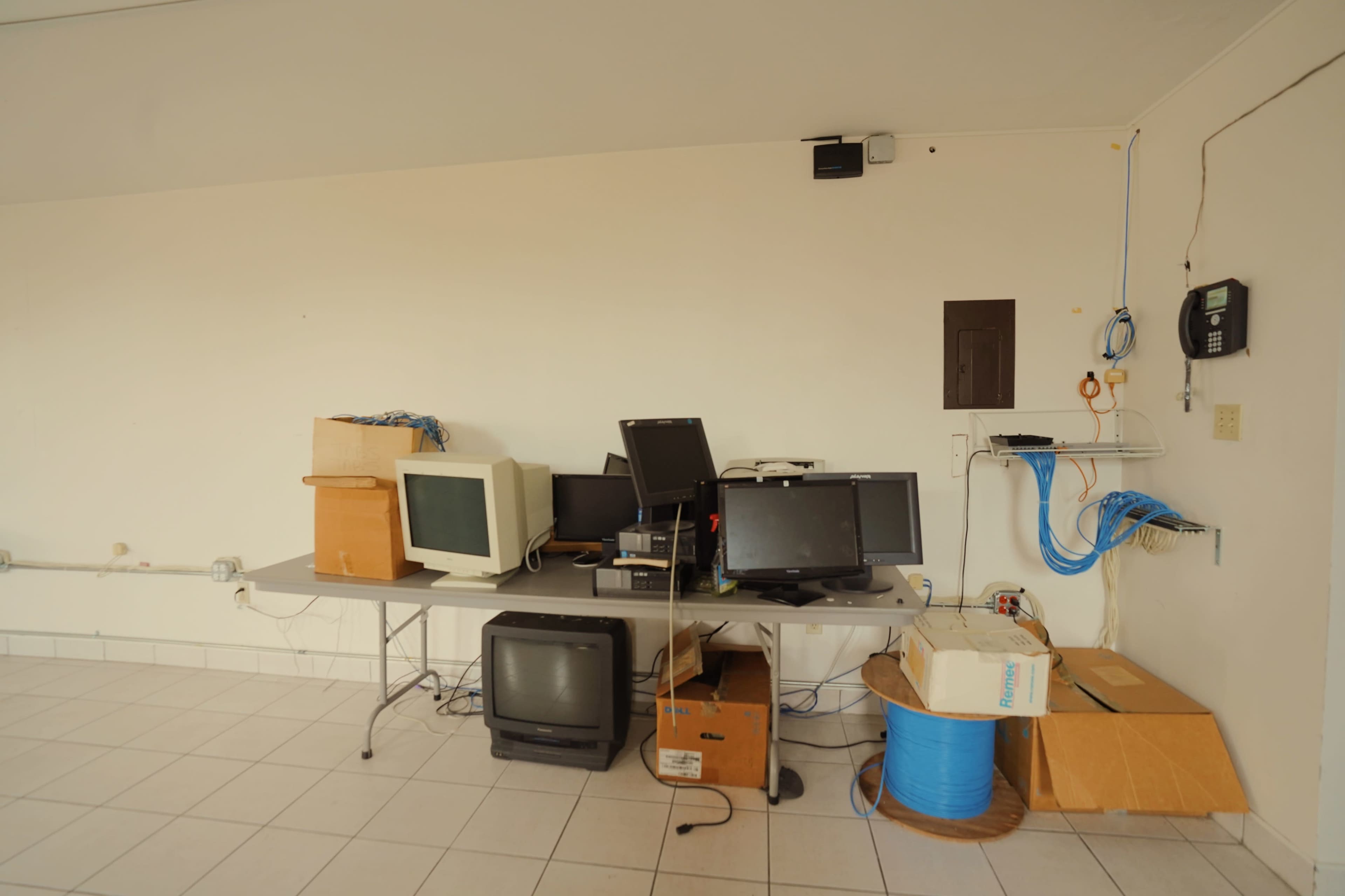 A table is lined with various old computer monitors, a television, and cardboard boxes in an empty room with tiled flooring.