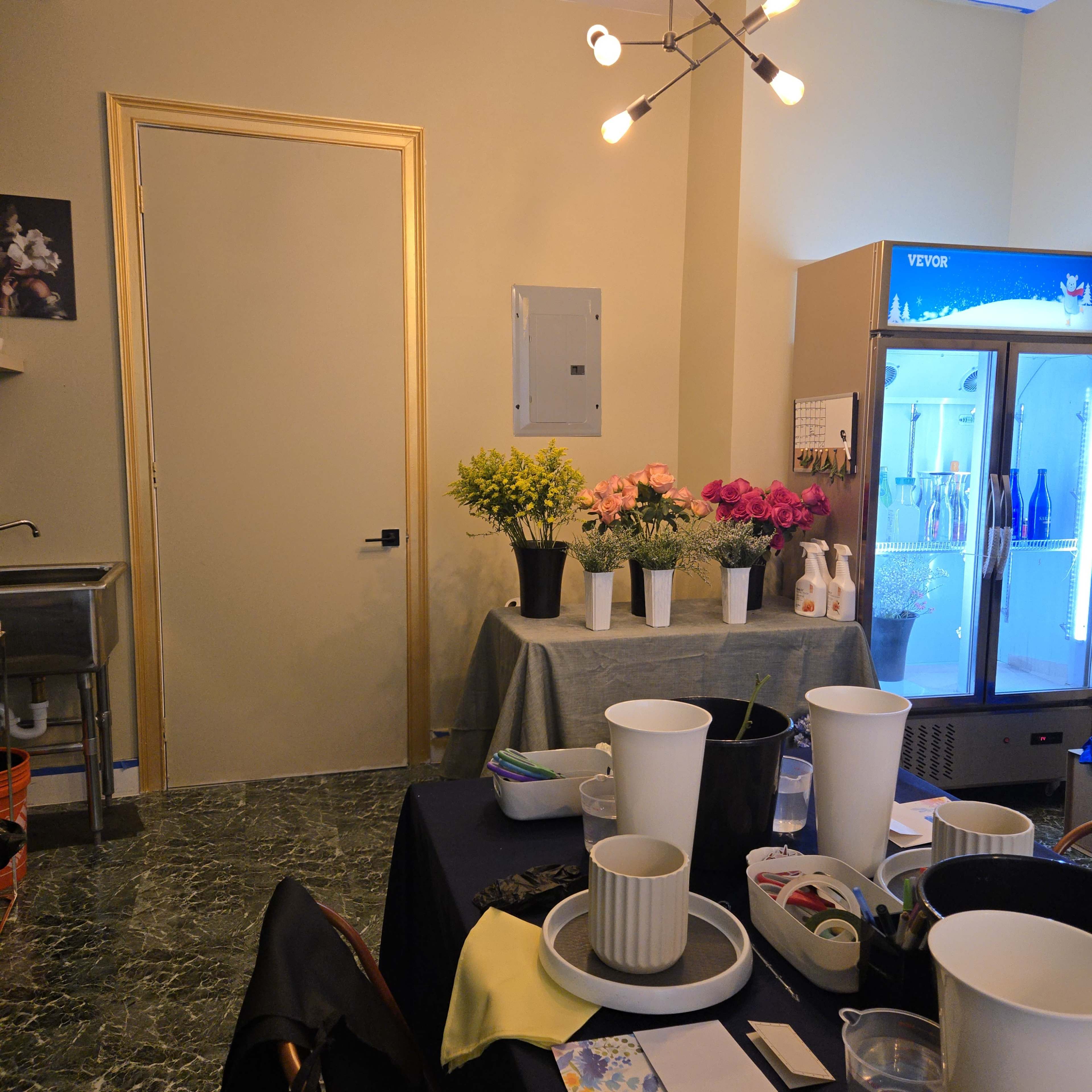 The image shows a room with a sink, a table covered with several potted flowers, and a refrigerator in the corner.