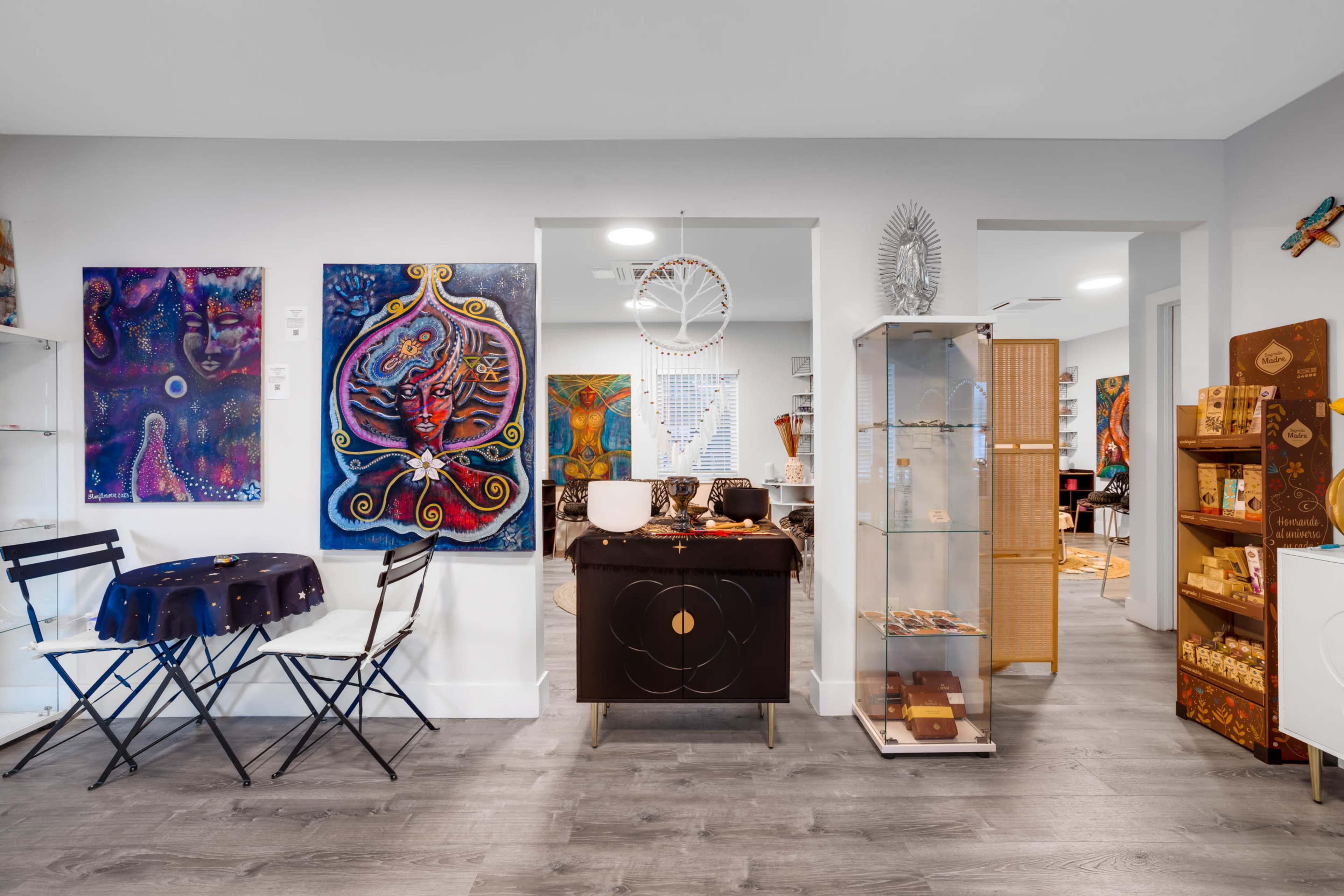 The image shows a colorful interior space featuring various artworks on the walls, a glass display case, and a small dining area with metal folding chairs and a round table.