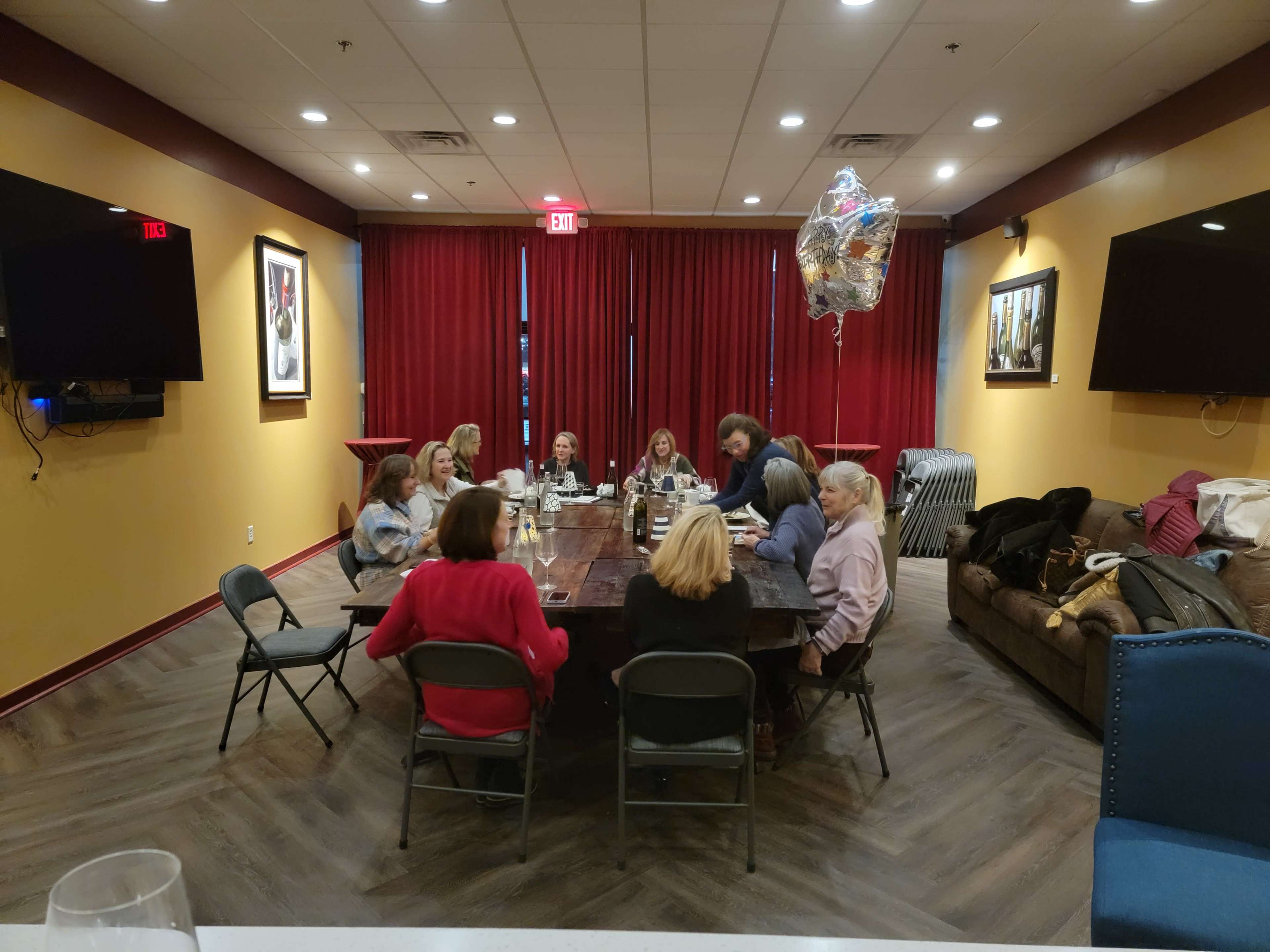 A group of people is seated around a large wooden table in a room with red curtains and light-colored walls.