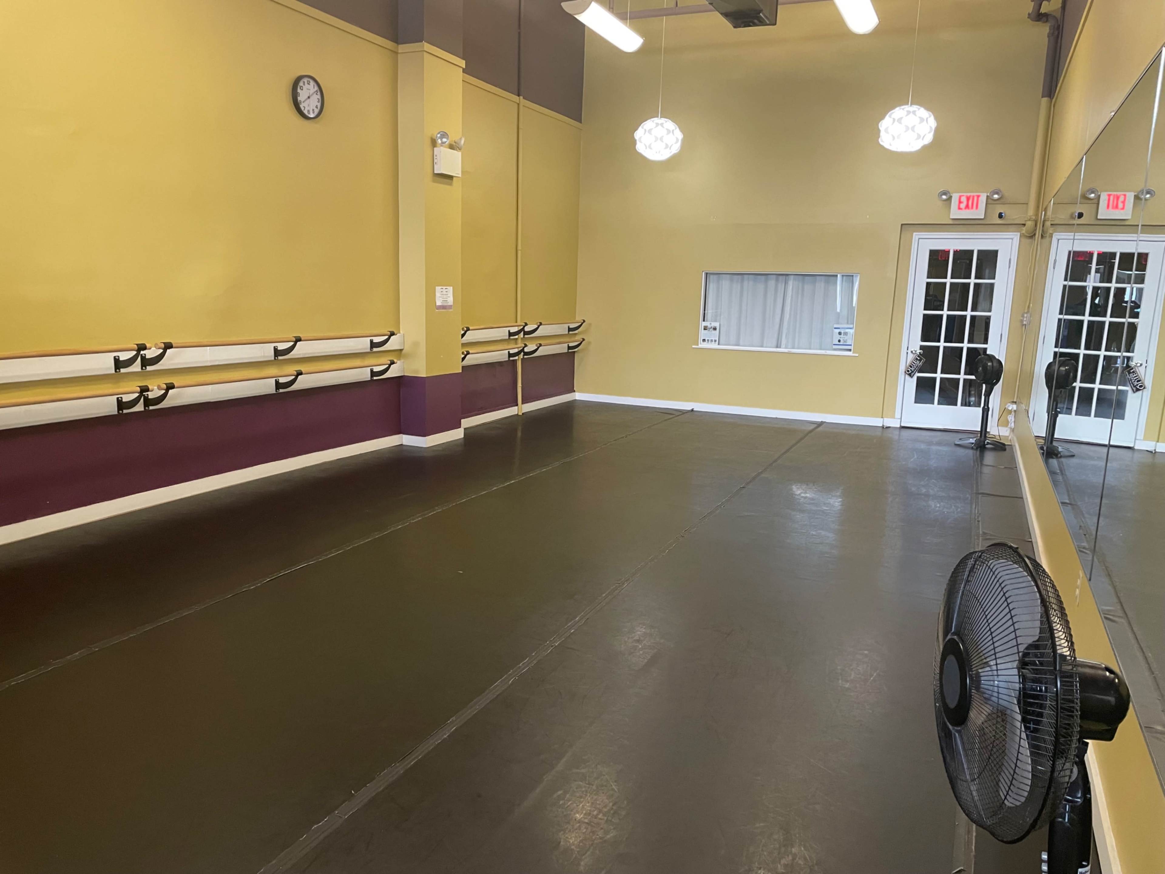 The image shows a dance studio with yellow walls, mirrors, ballet barres, and a ceiling fan in one corner.