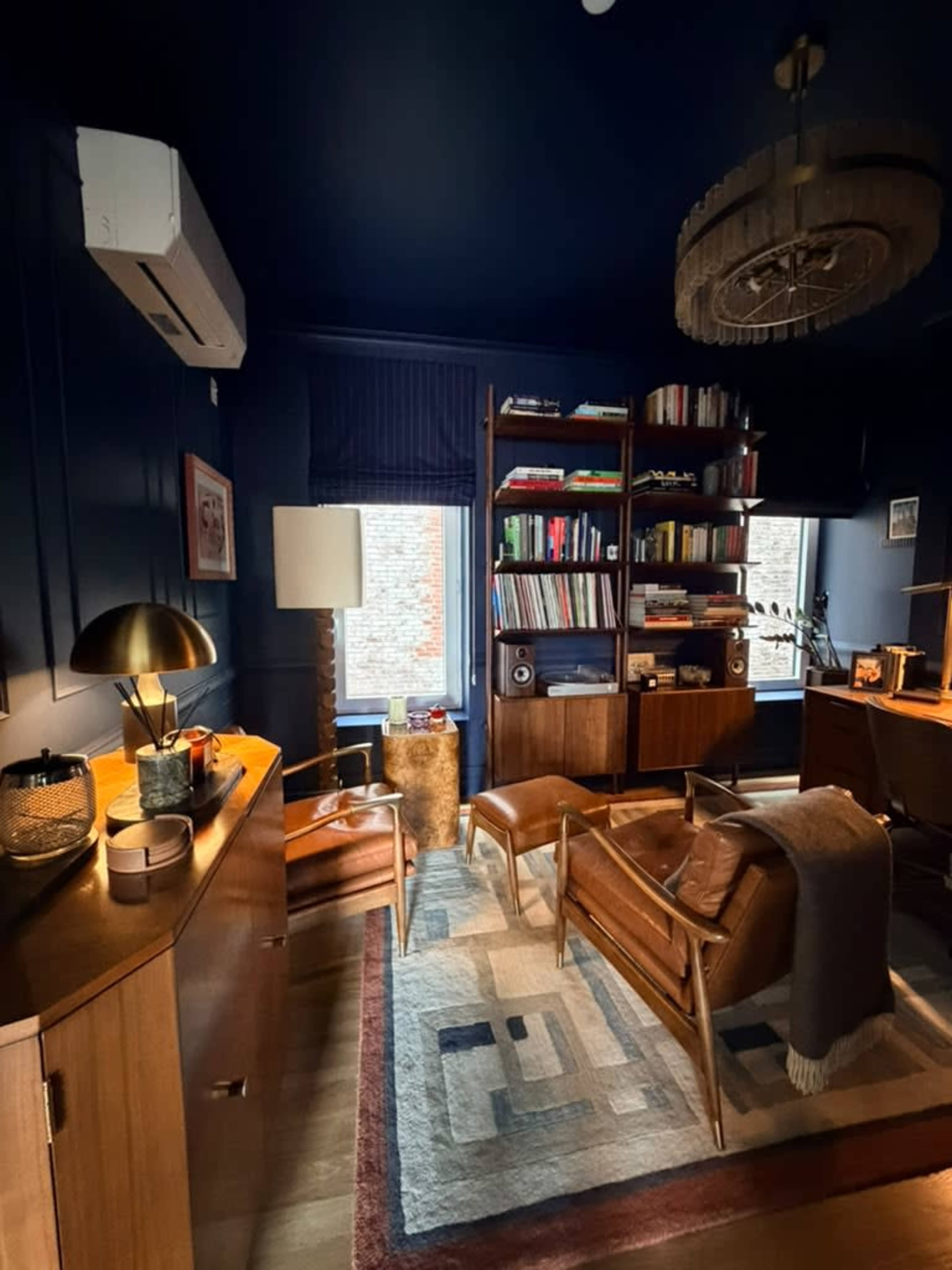 The image shows a stylish office space with dark blue walls, wooden furniture, a bookshelf filled with books, and two armchairs arranged around a small table.