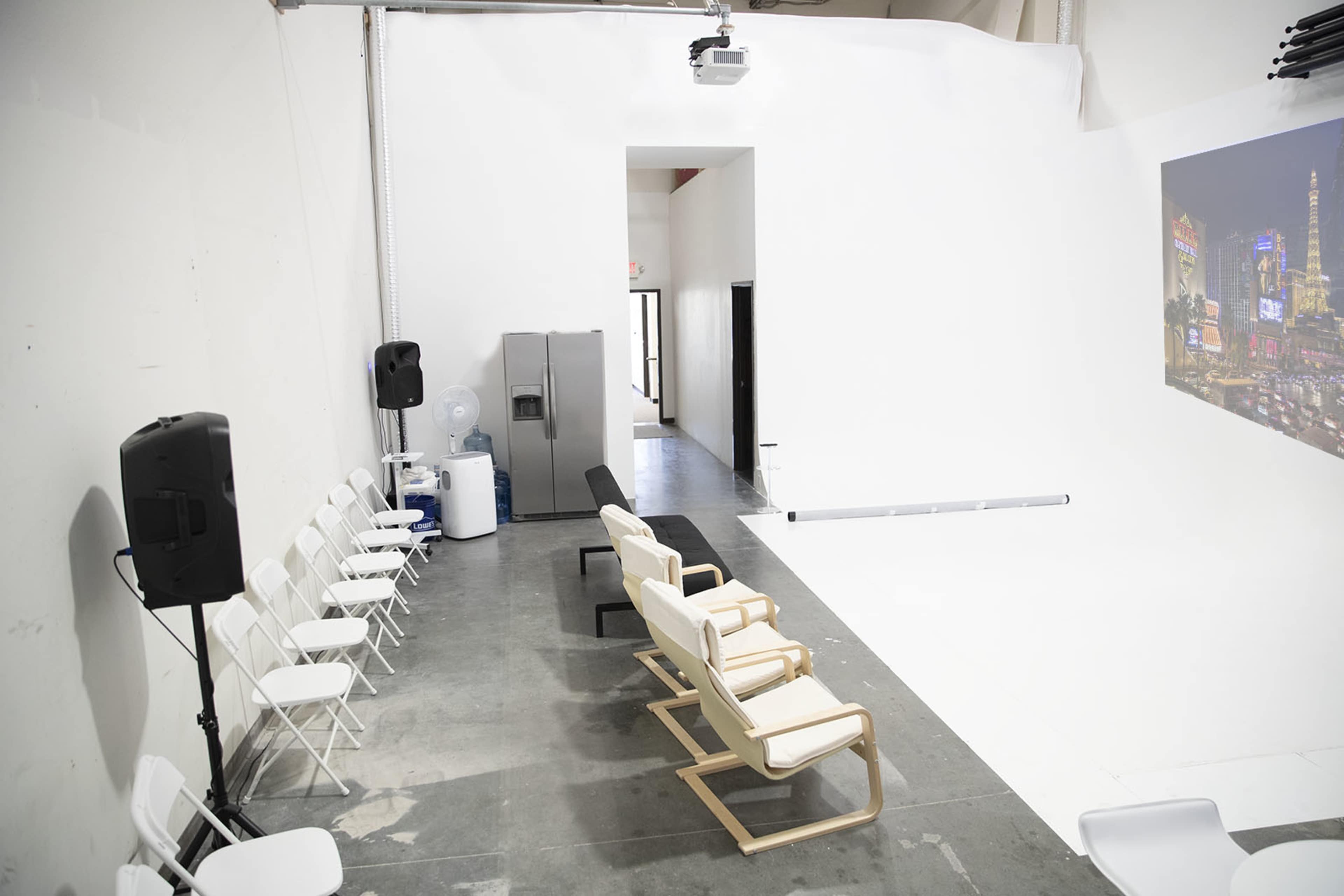 A spacious studio featuring a row of white chairs, a black bench, and a projection of a cityscape on a white wall.