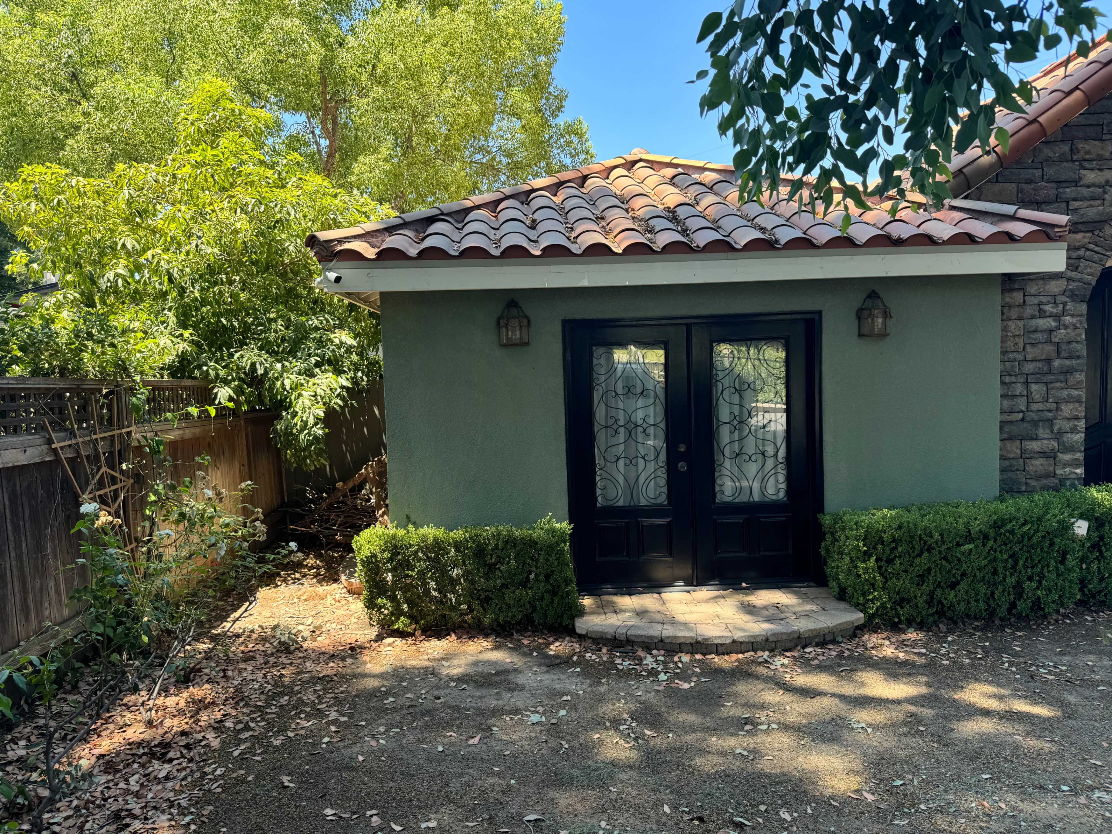 A small, green building with a tiled roof and double black doors, surrounded by greenery and a gravel pathway.