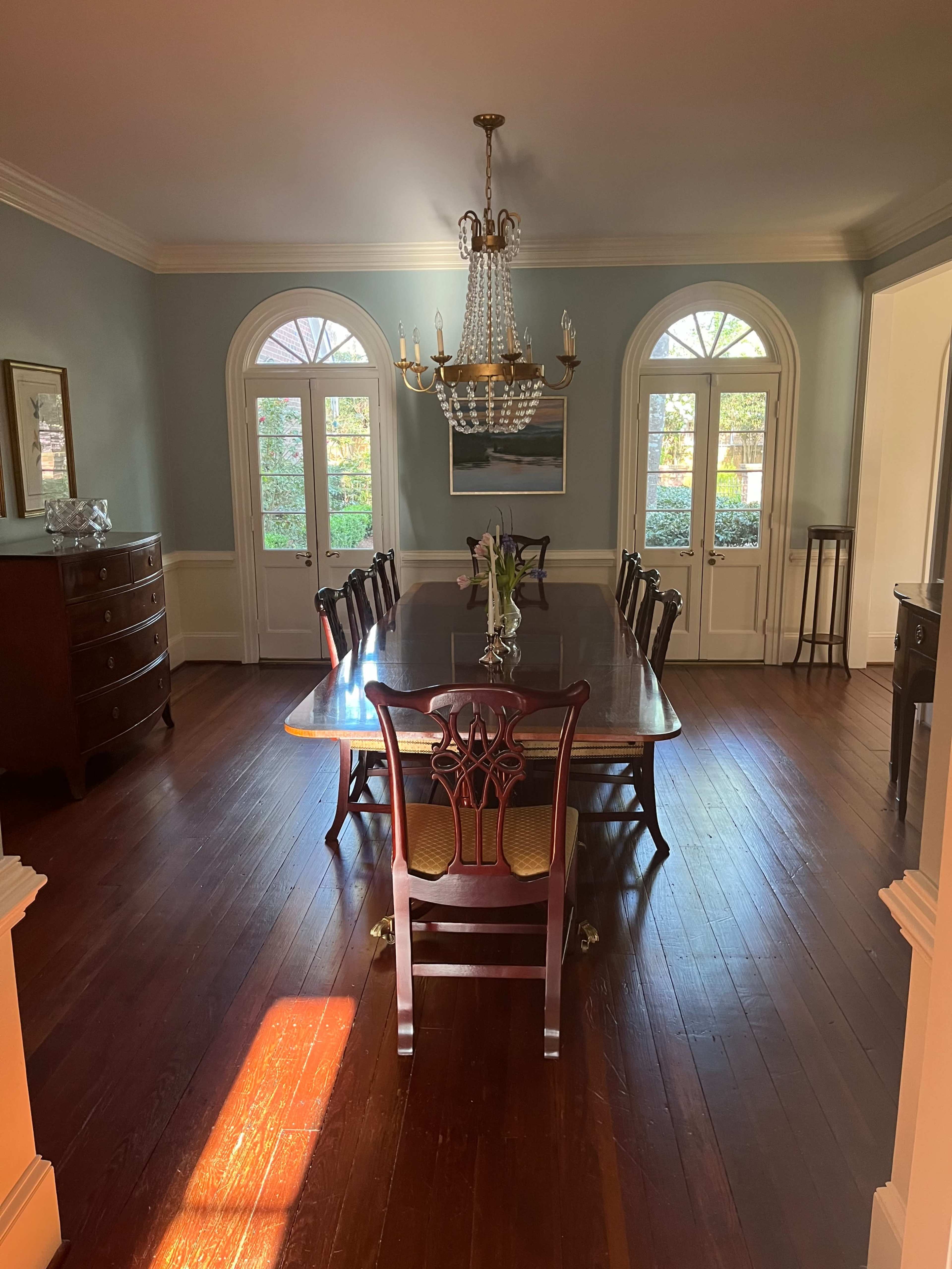 The image shows a spacious dining room with a large wooden table surrounded by chairs, a chandelier above, and large windows letting in natural light.