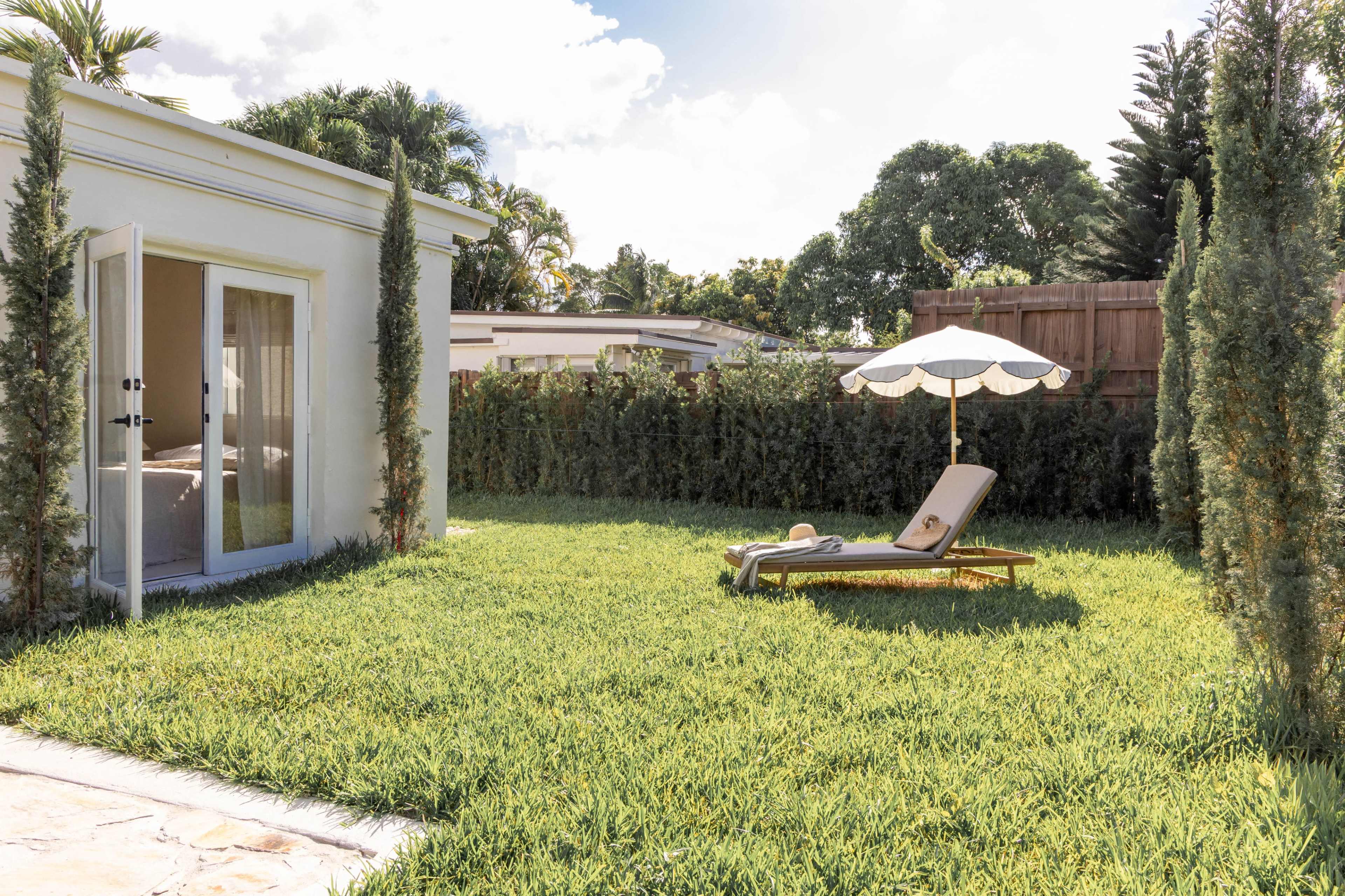 A lounge chair sits under a white umbrella in a grassy backyard next to a small house with open French doors.
