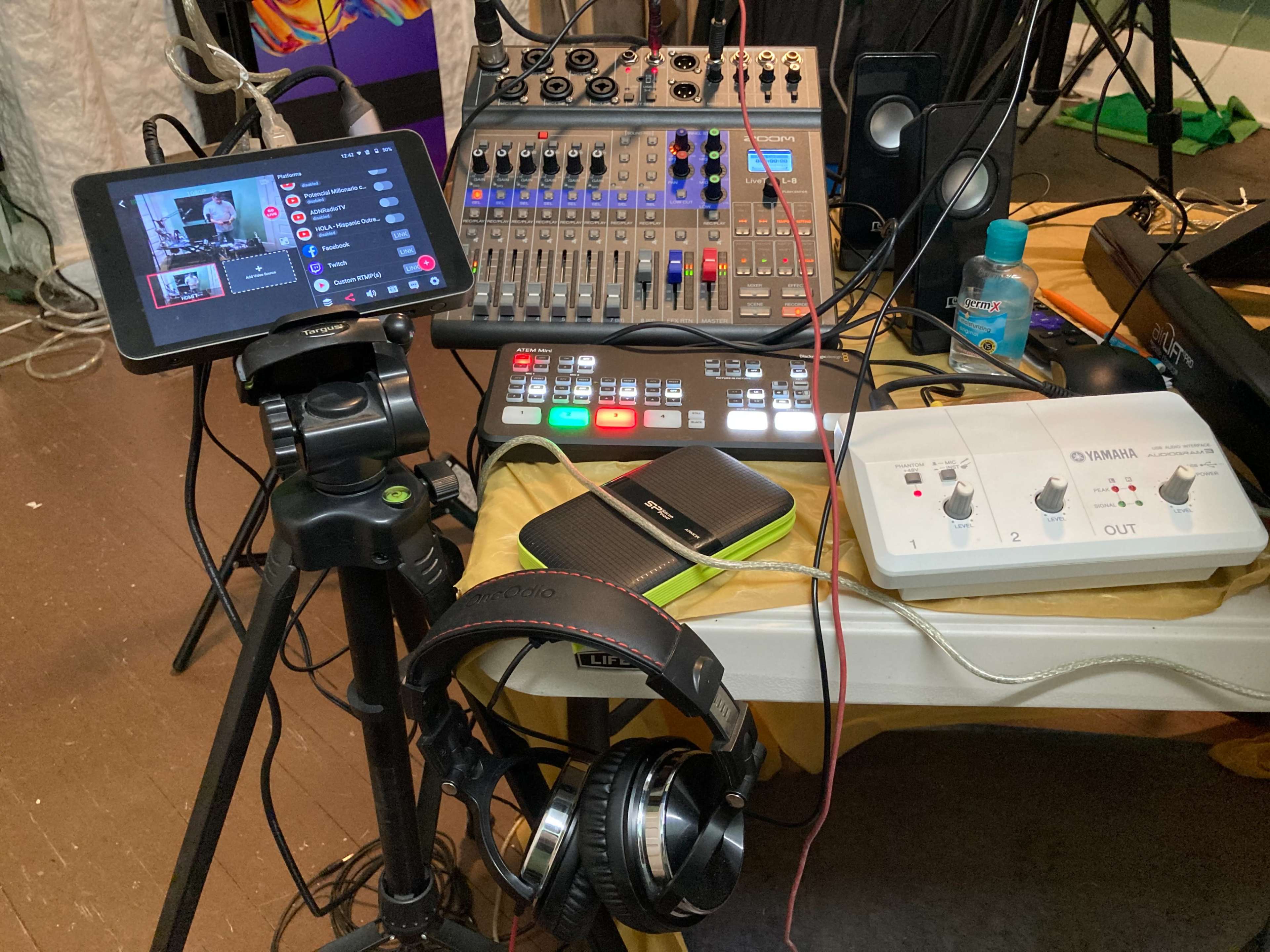 A recording setup featuring a tablet on a tripod, a multi-channel audio mixer, a smartphone, a pair of headphones, and an audio interface on a table.