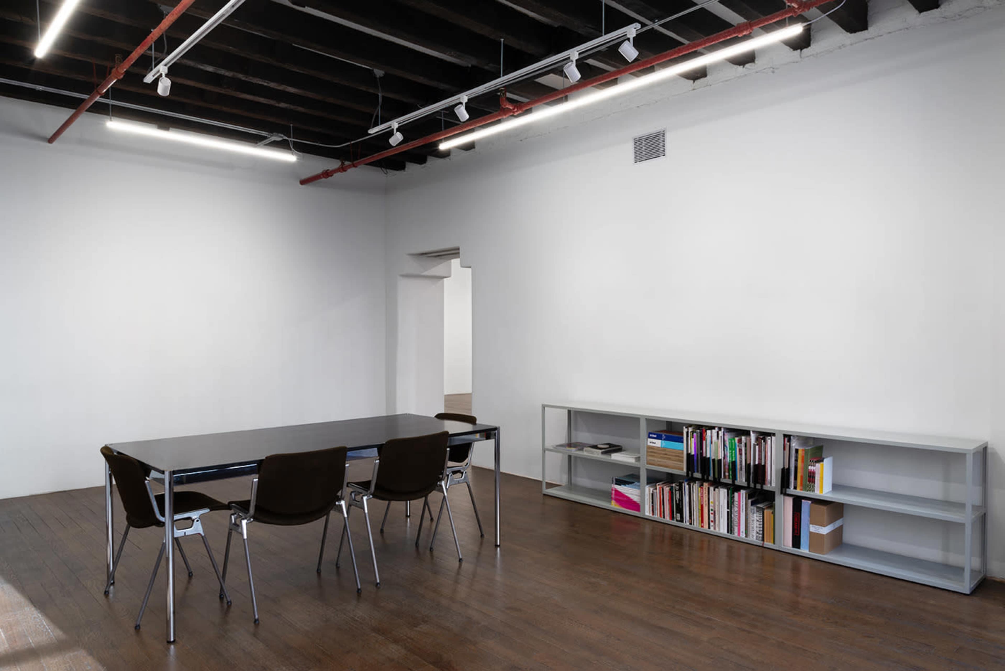 The image shows a minimalist office space with a large black table surrounded by four chairs, and a bookshelf along one wall filled with books.