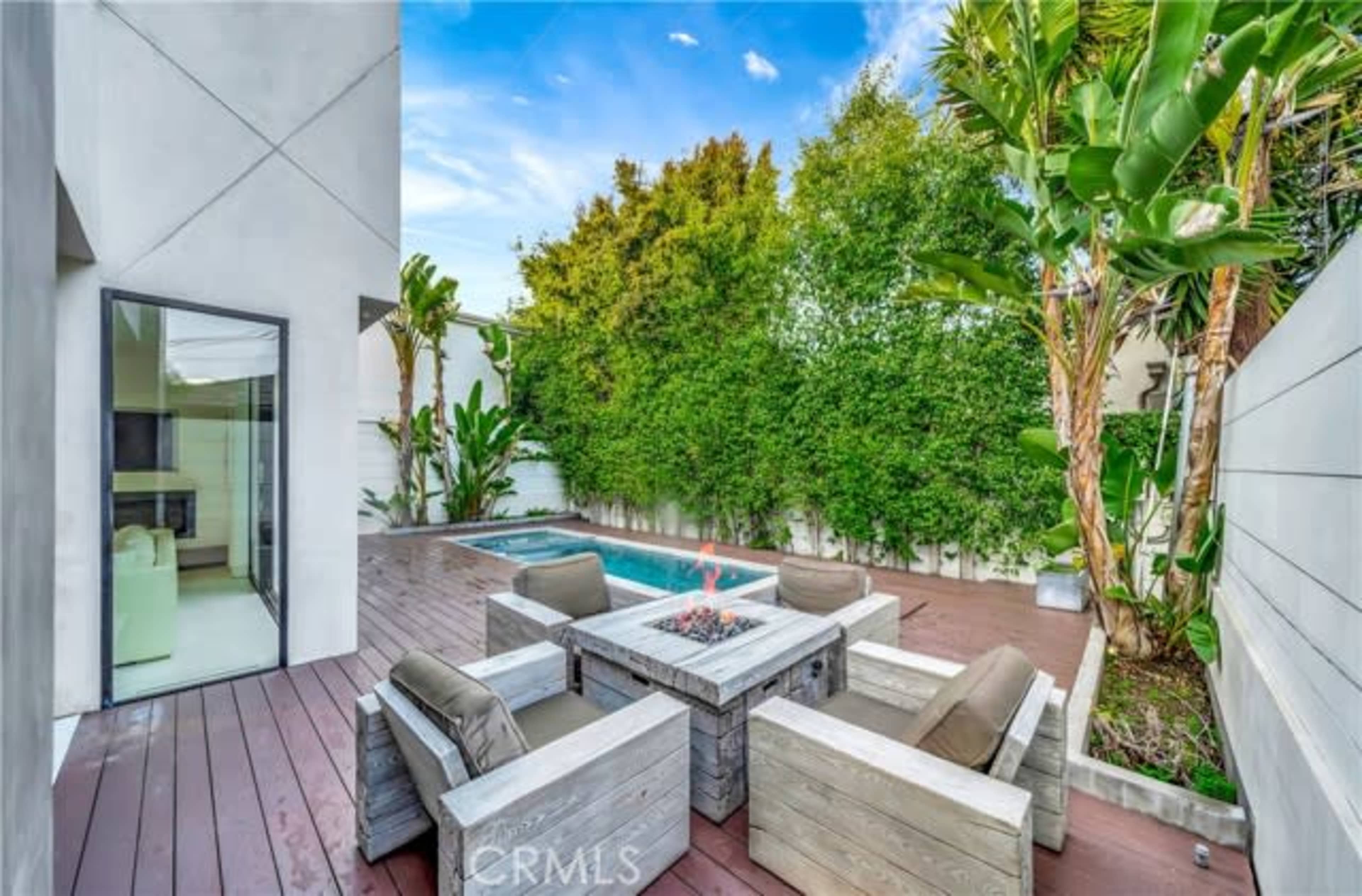 The image shows a modern patio area featuring a fire pit surrounded by seating, with a pool and lush greenery in the background.