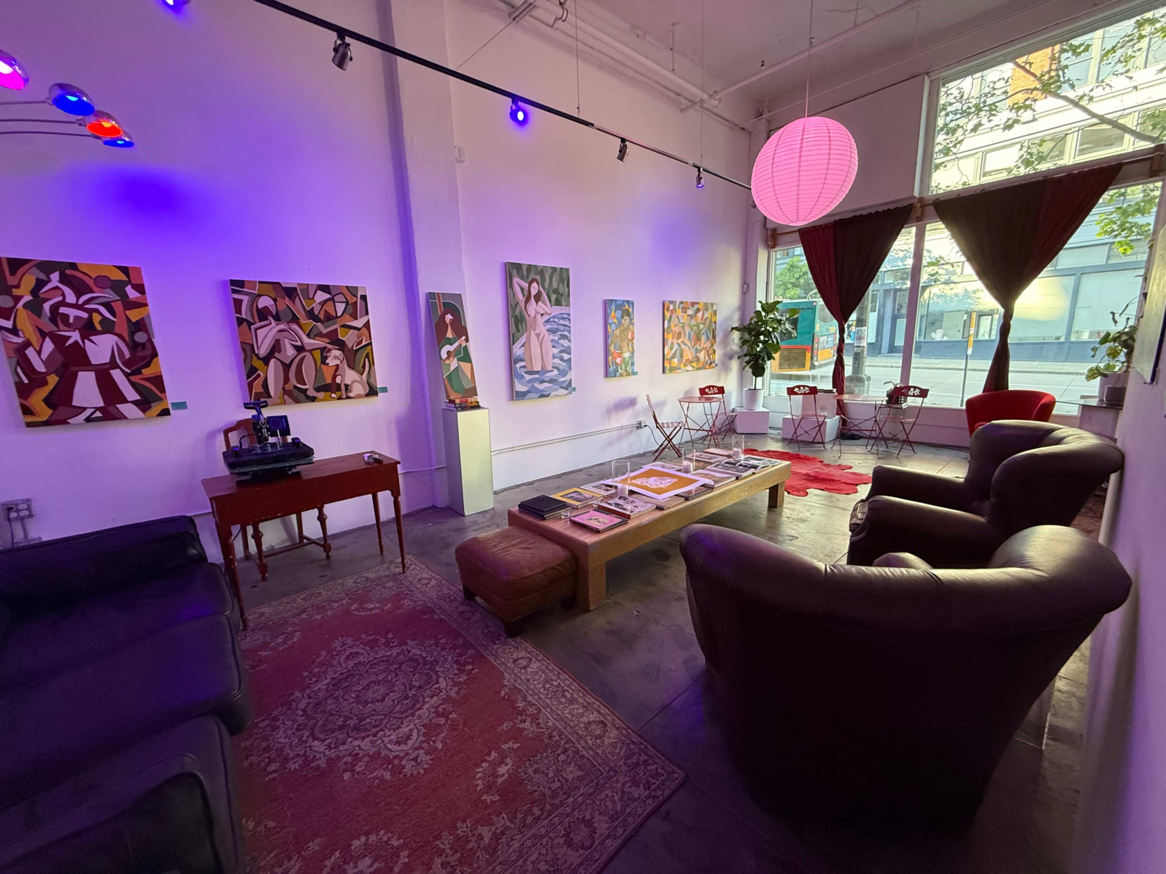 The image shows an art gallery with colorful paintings on the walls, a low table in the center, and seating areas featuring two armchairs and a couch.