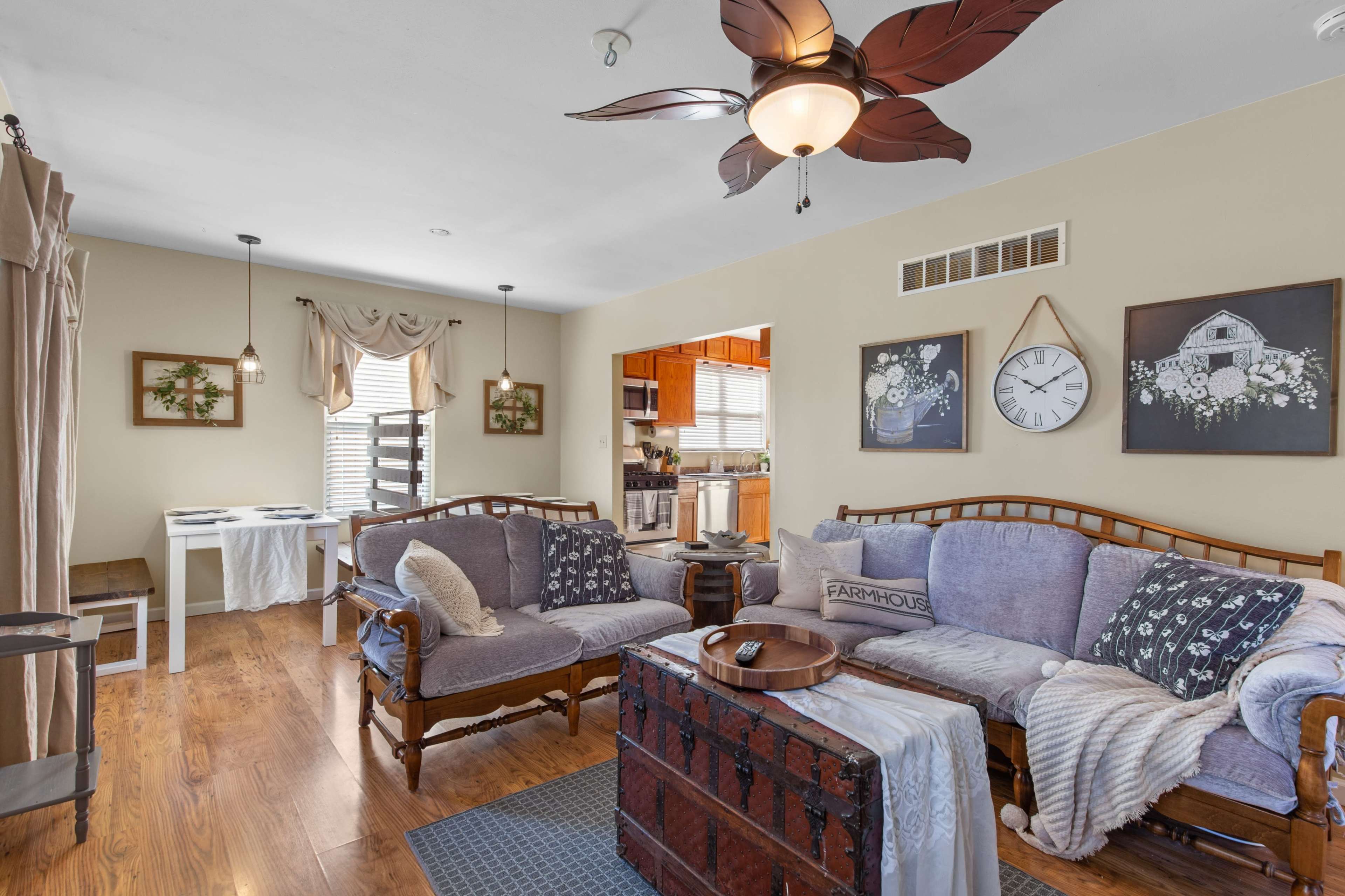 The image shows a cozy living room featuring a pair of wooden couches, a central trunk table, and decor including wall art, a clock, and a dining area visible in the background.