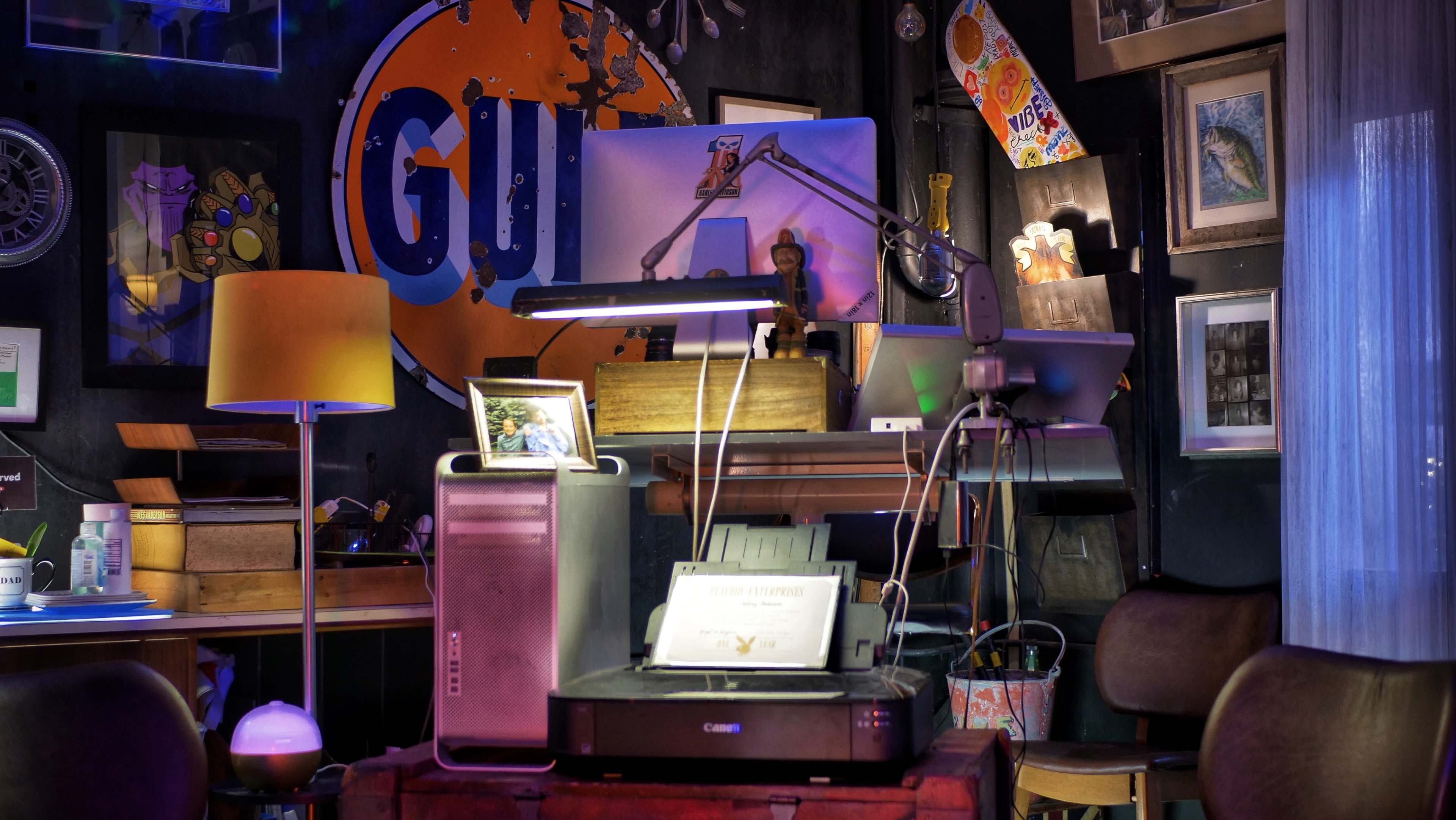 A cluttered workspace featuring a computer setup, a printer, various artworks on the walls, and a lamp, all in a room with a large "GUT" logo painted on the wall.