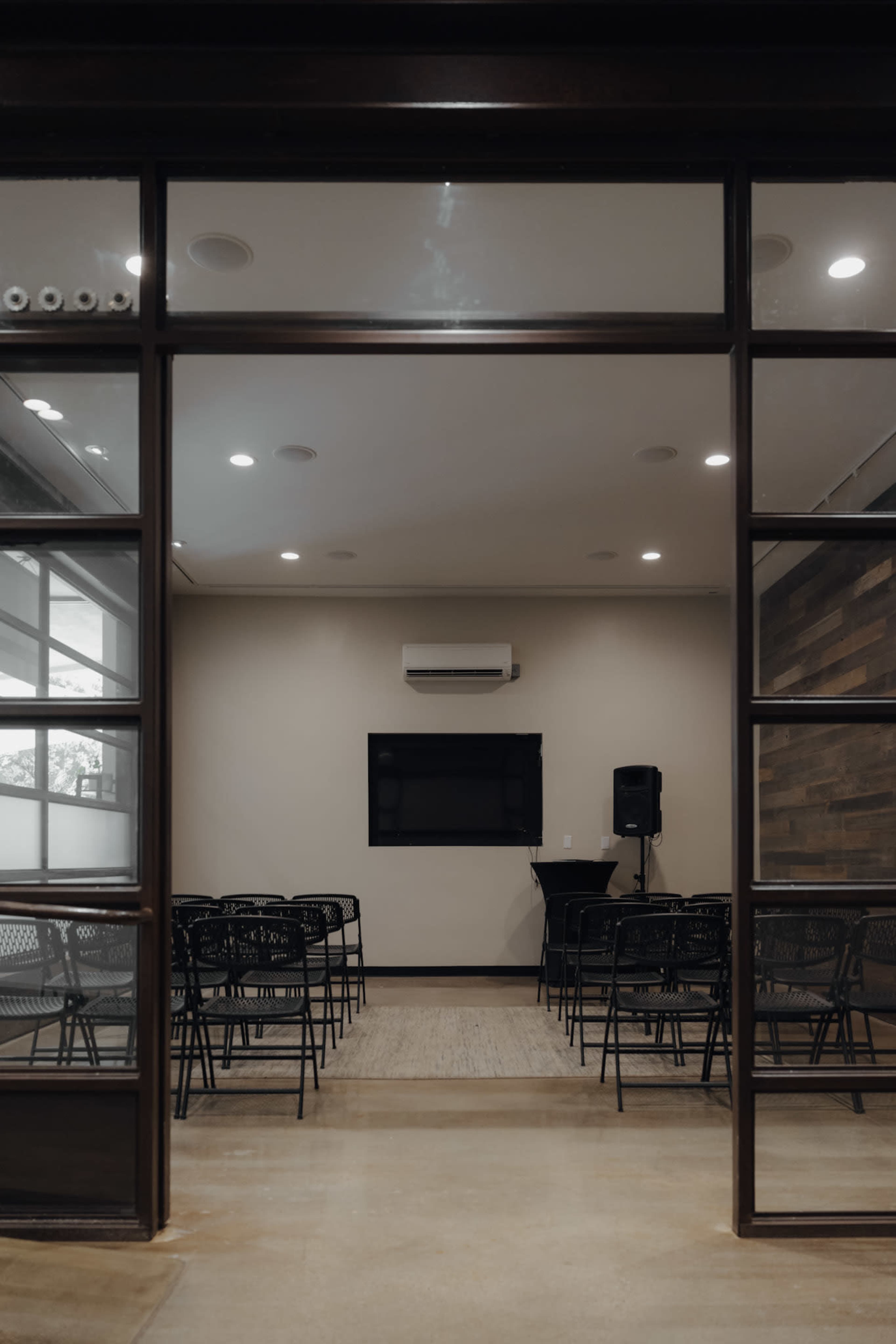A conference room is seen through glass doors, featuring multiple rows of black chairs facing a wall-mounted TV and a small podium.