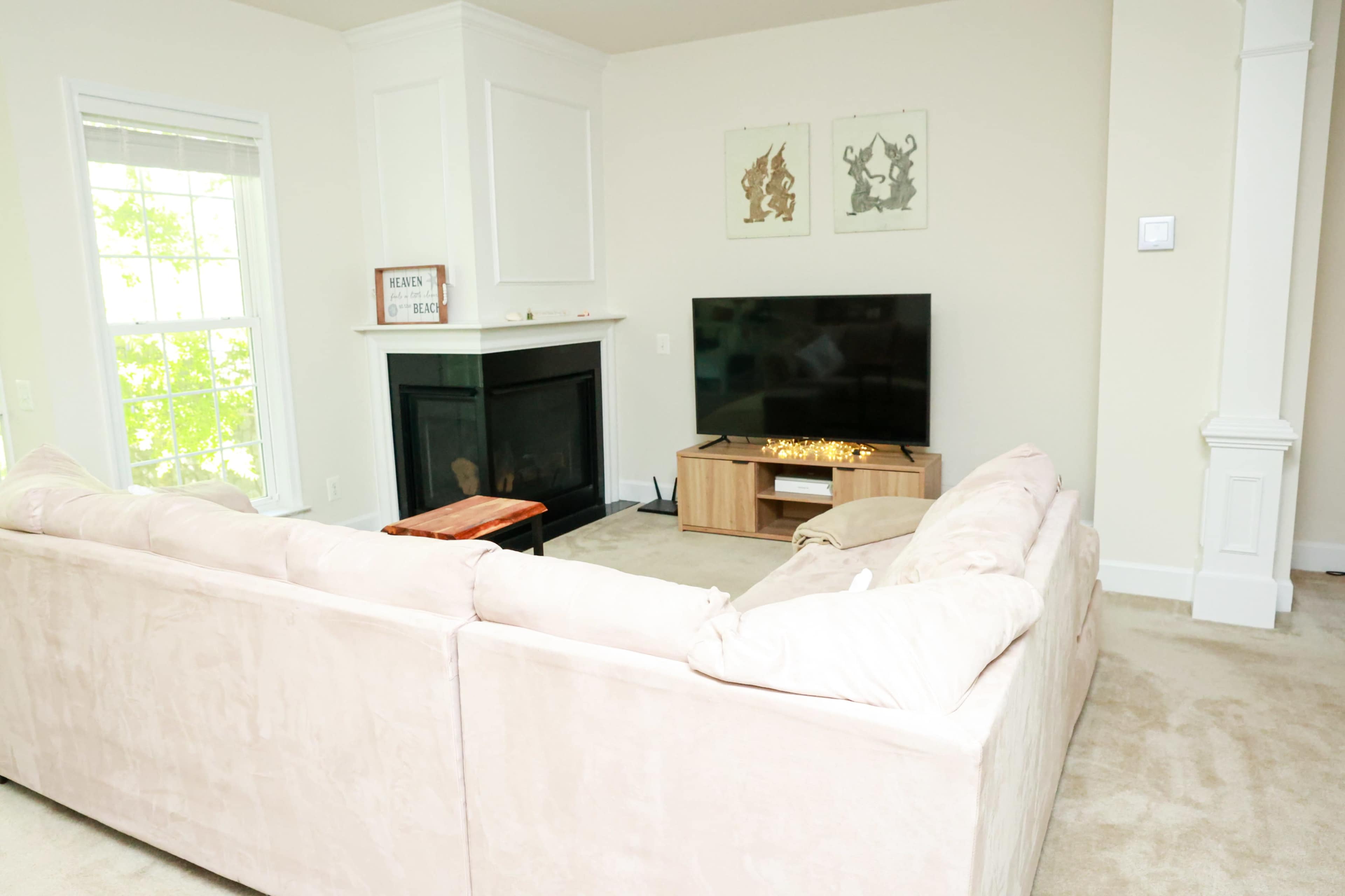 A cozy living room features a beige sectional sofa, a wall-mounted television, and a fireplace, with light streaming in through the window.