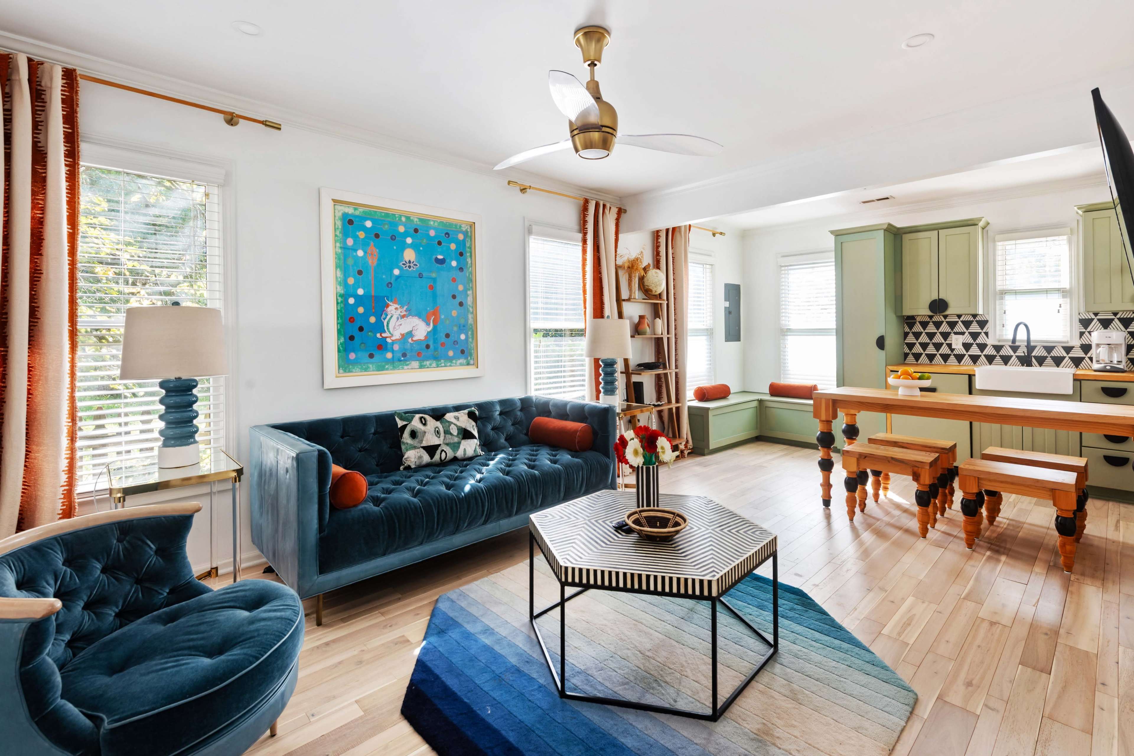 The image shows a bright living space with a blue velvet sofa, a round coffee table, and a dining area featuring green cabinets and a patterned backsplash.