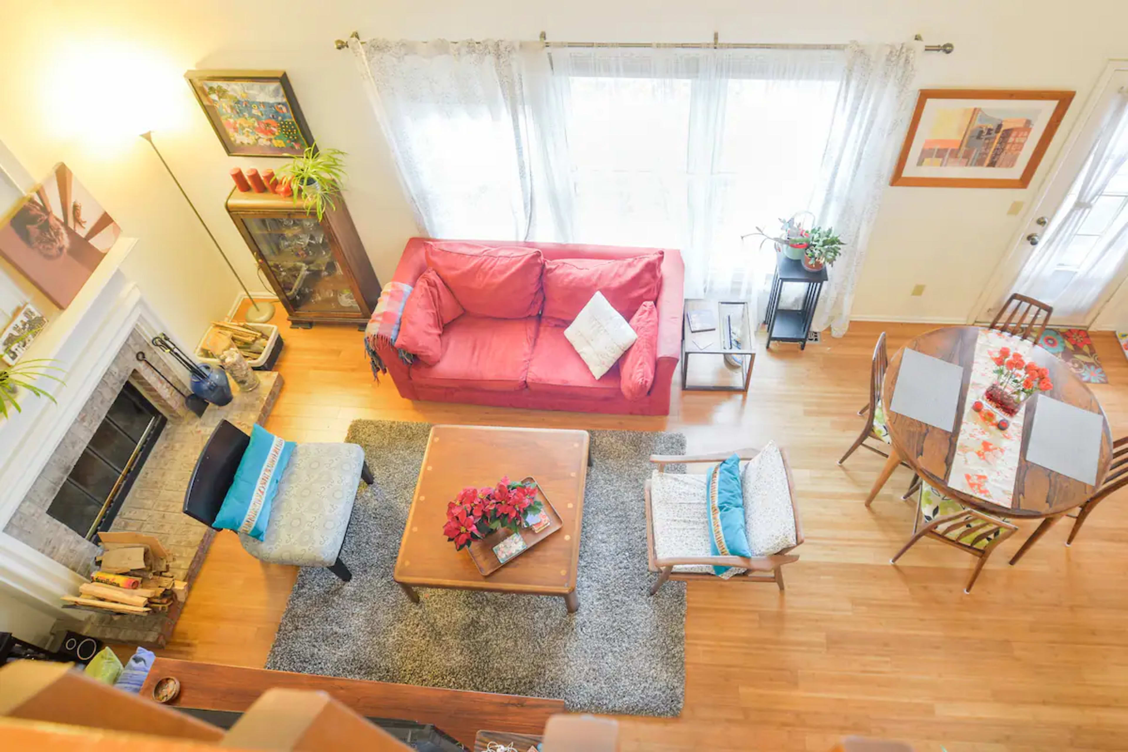 A bright living room with a red couch, a coffee table, an area rug, and a dining table set for four, surrounded by large windows.