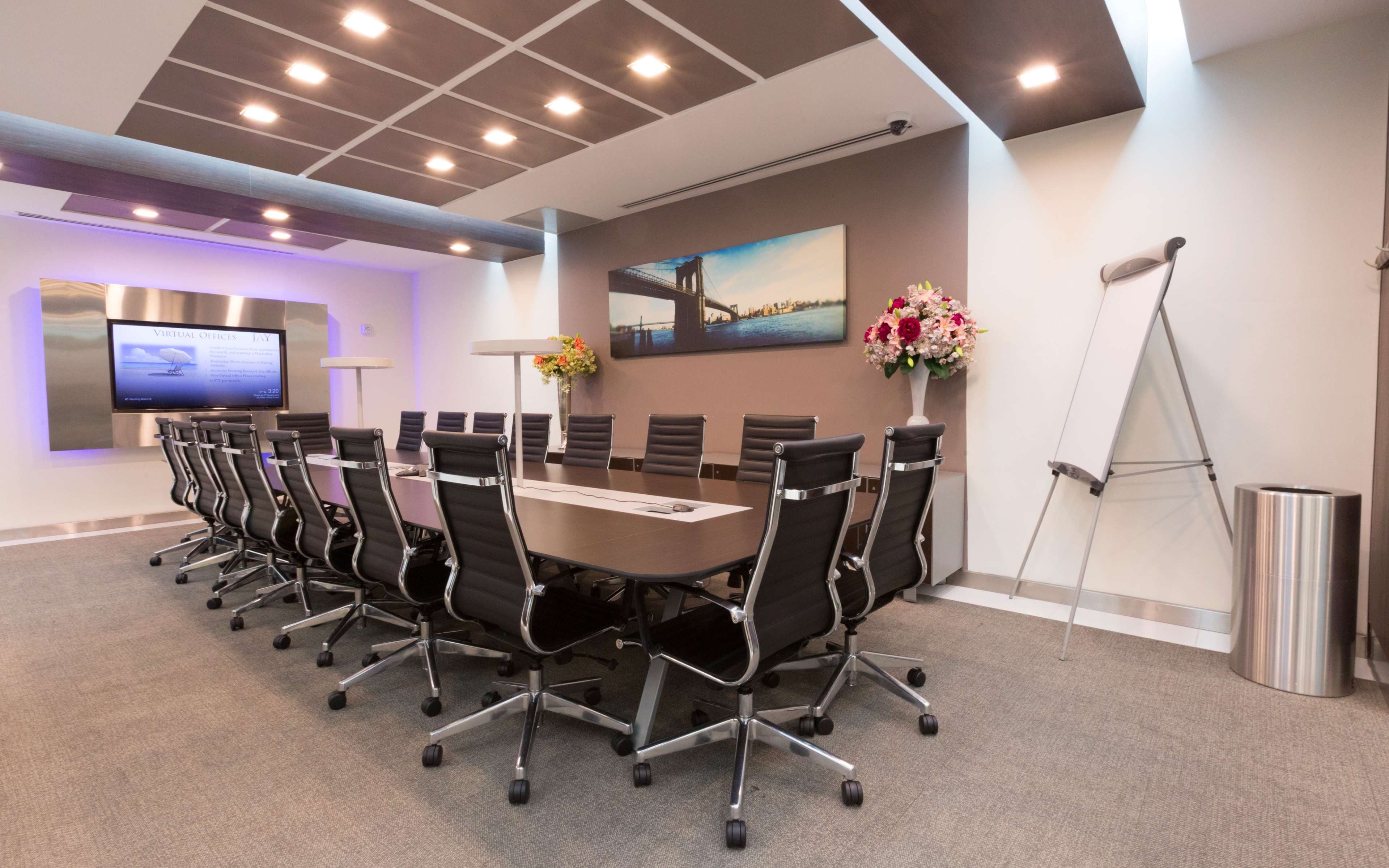 A modern conference room features a large table surrounded by black ergonomic chairs, a mounted screen, and flowers arranged near a whiteboard.