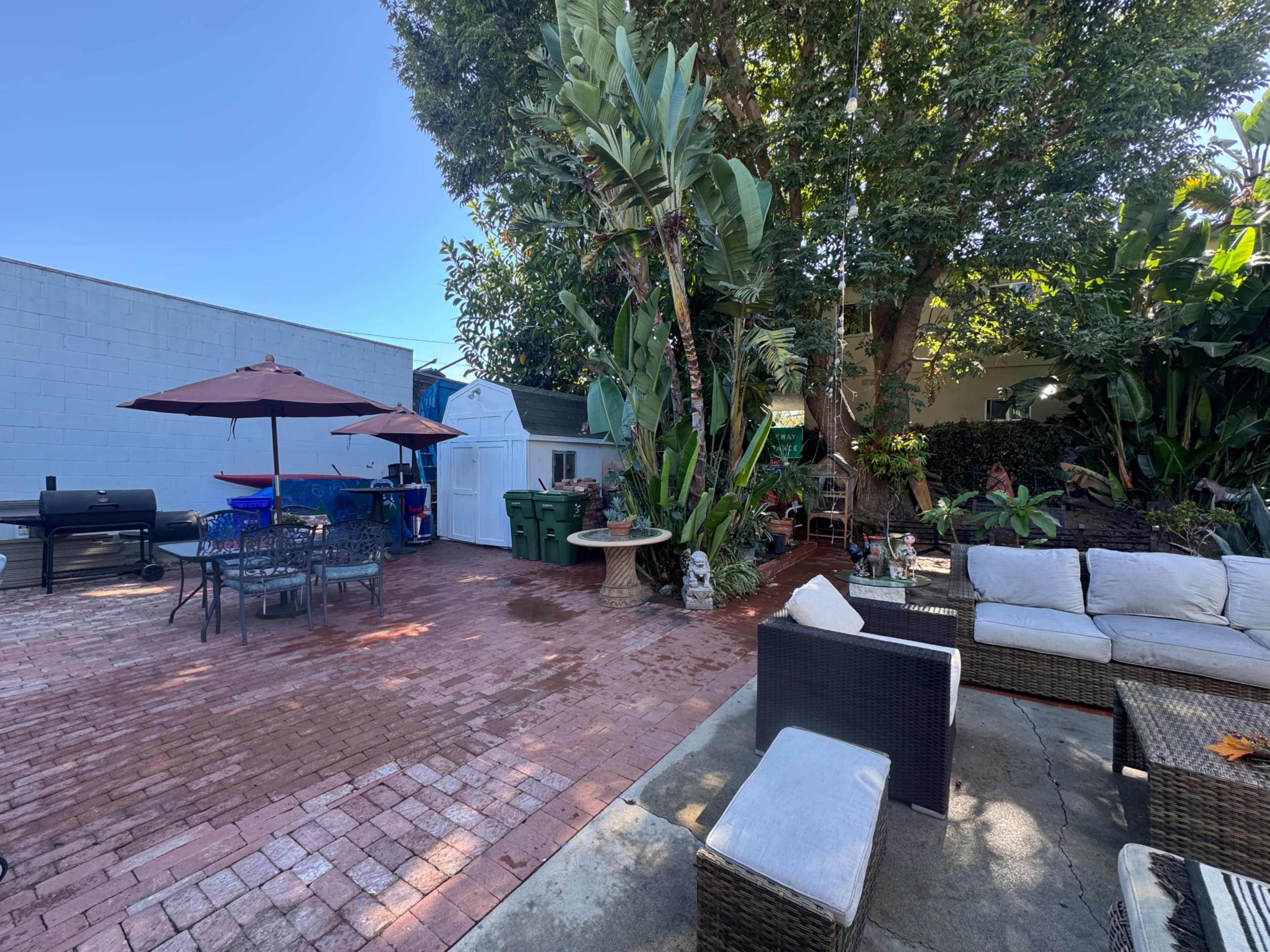 The image features a backyard patio area with brick flooring, outdoor seating, a barbeque grill, and potted plants under a clear blue sky.