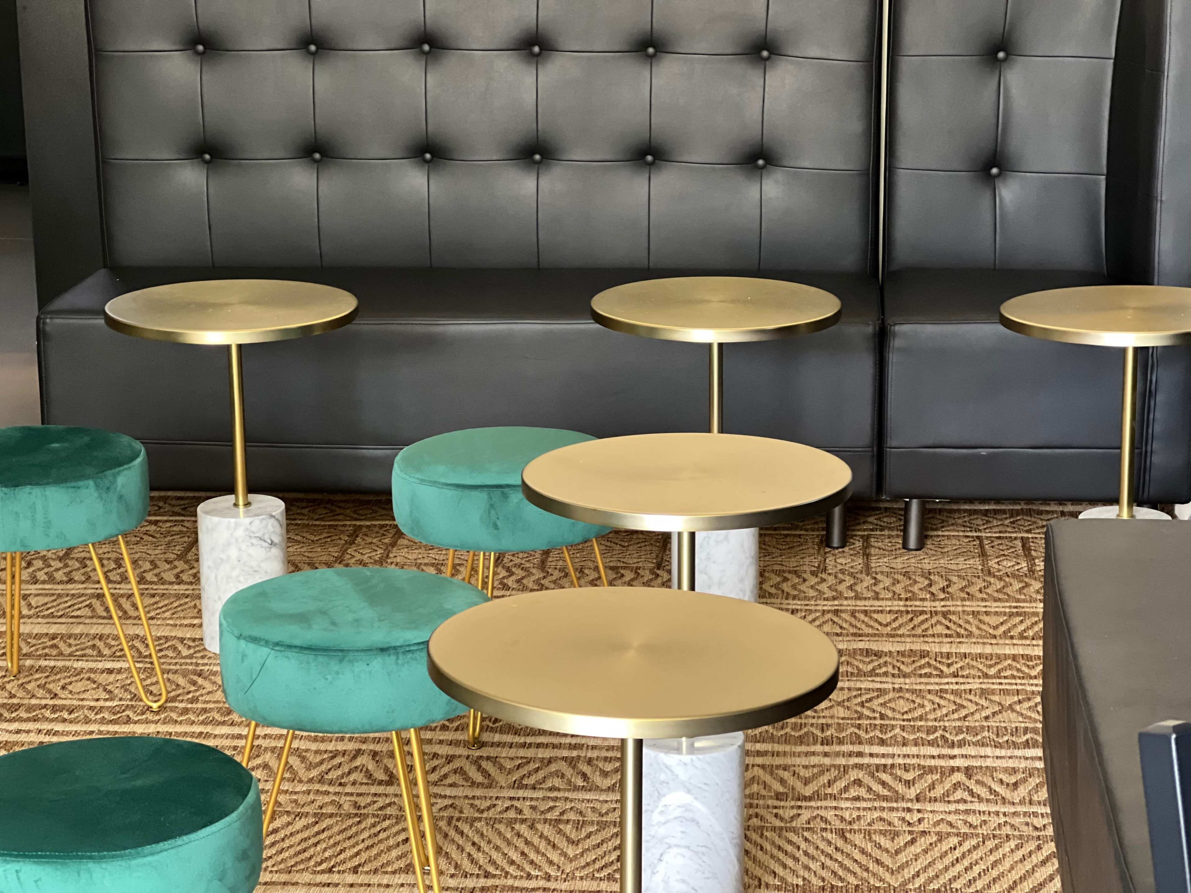 A modern seating area with a black tufted sofa and several small round tables with gold tops and green upholstered stools.