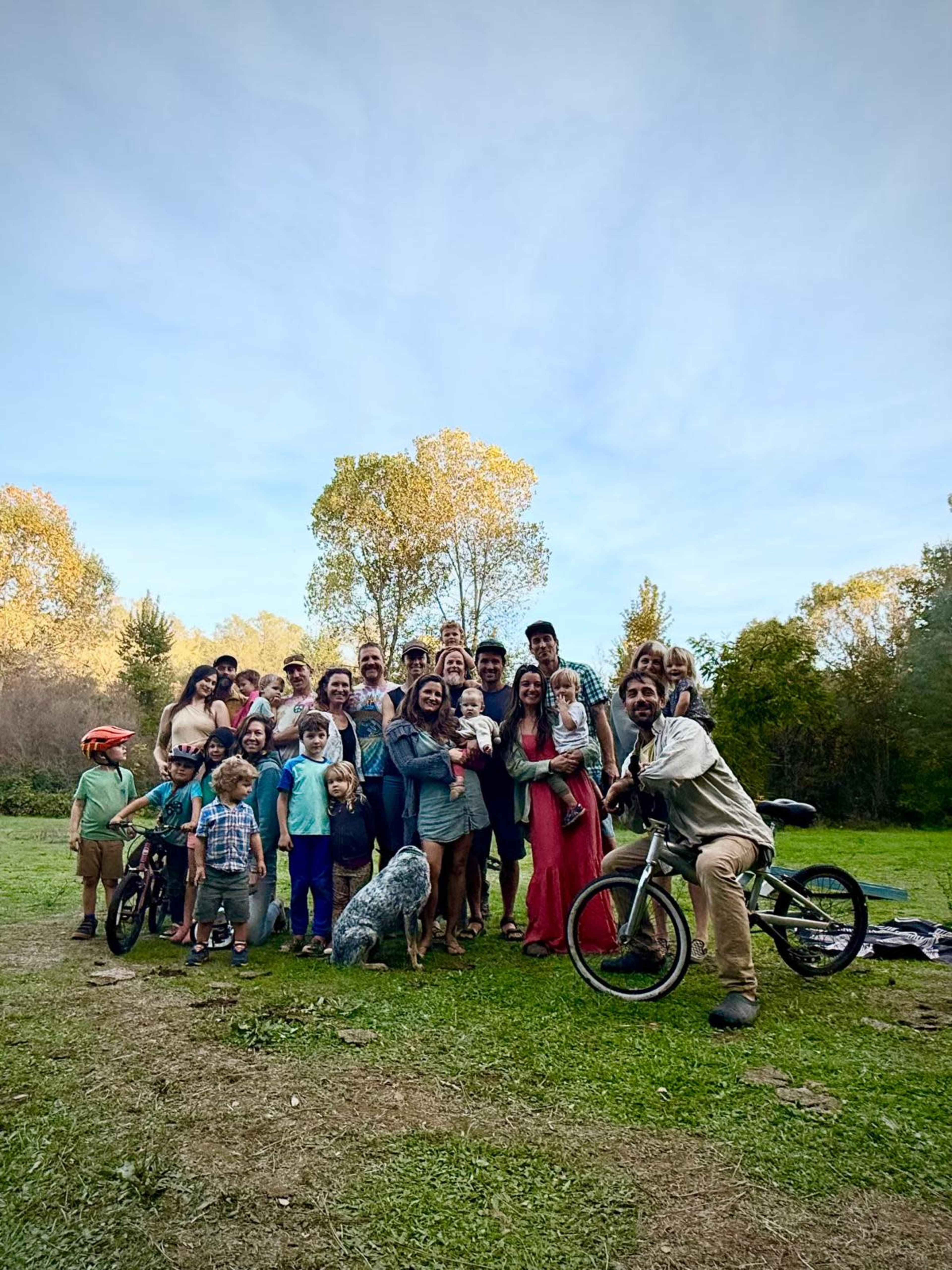 A large group of people, both adults and children, gather in a grassy area with trees in the background, along with several dogs and a bicycle.