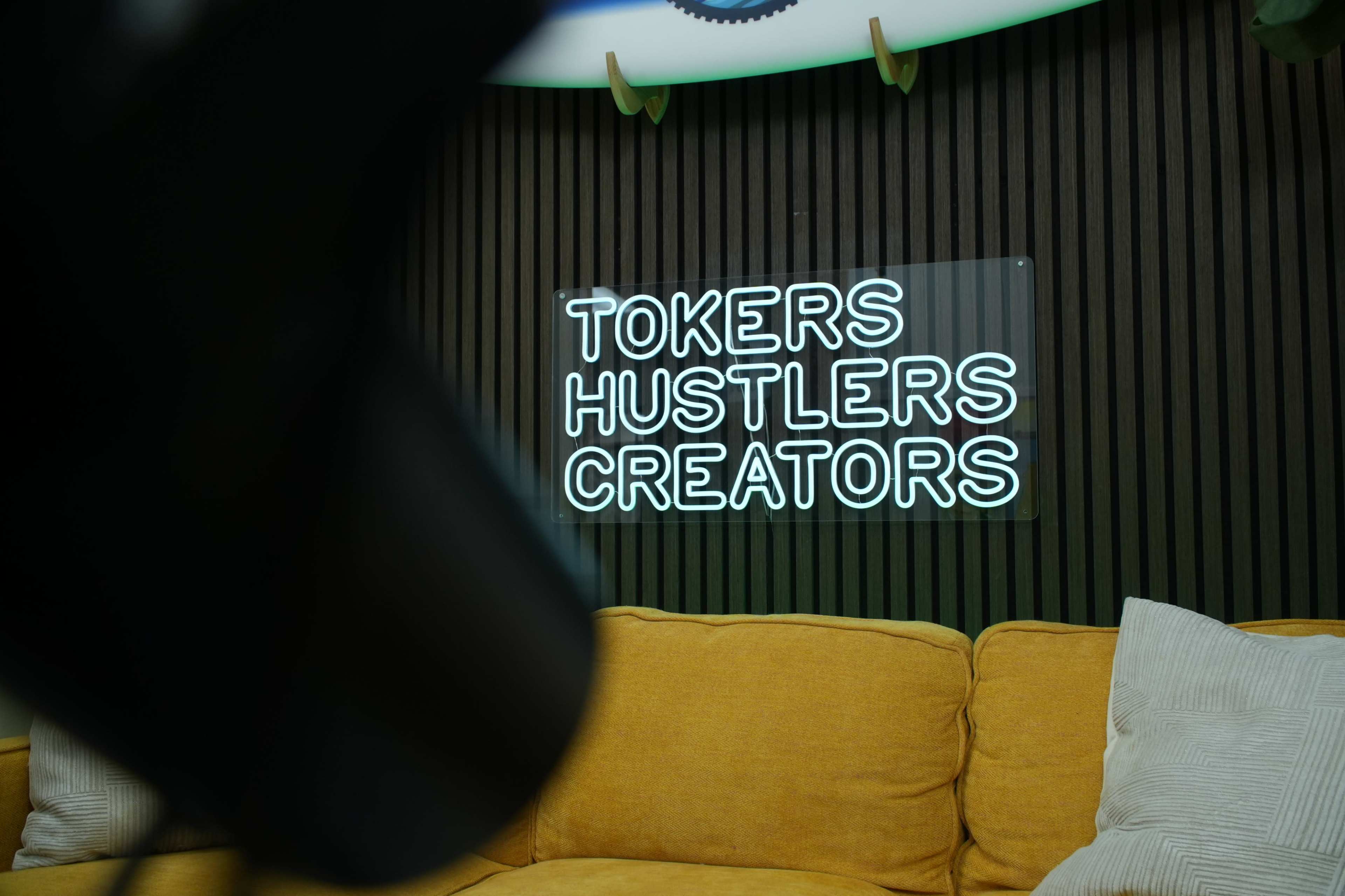 A neon sign that reads "TOKERS HUSTLERS CREATORS" is mounted on a textured wall above a yellow couch.