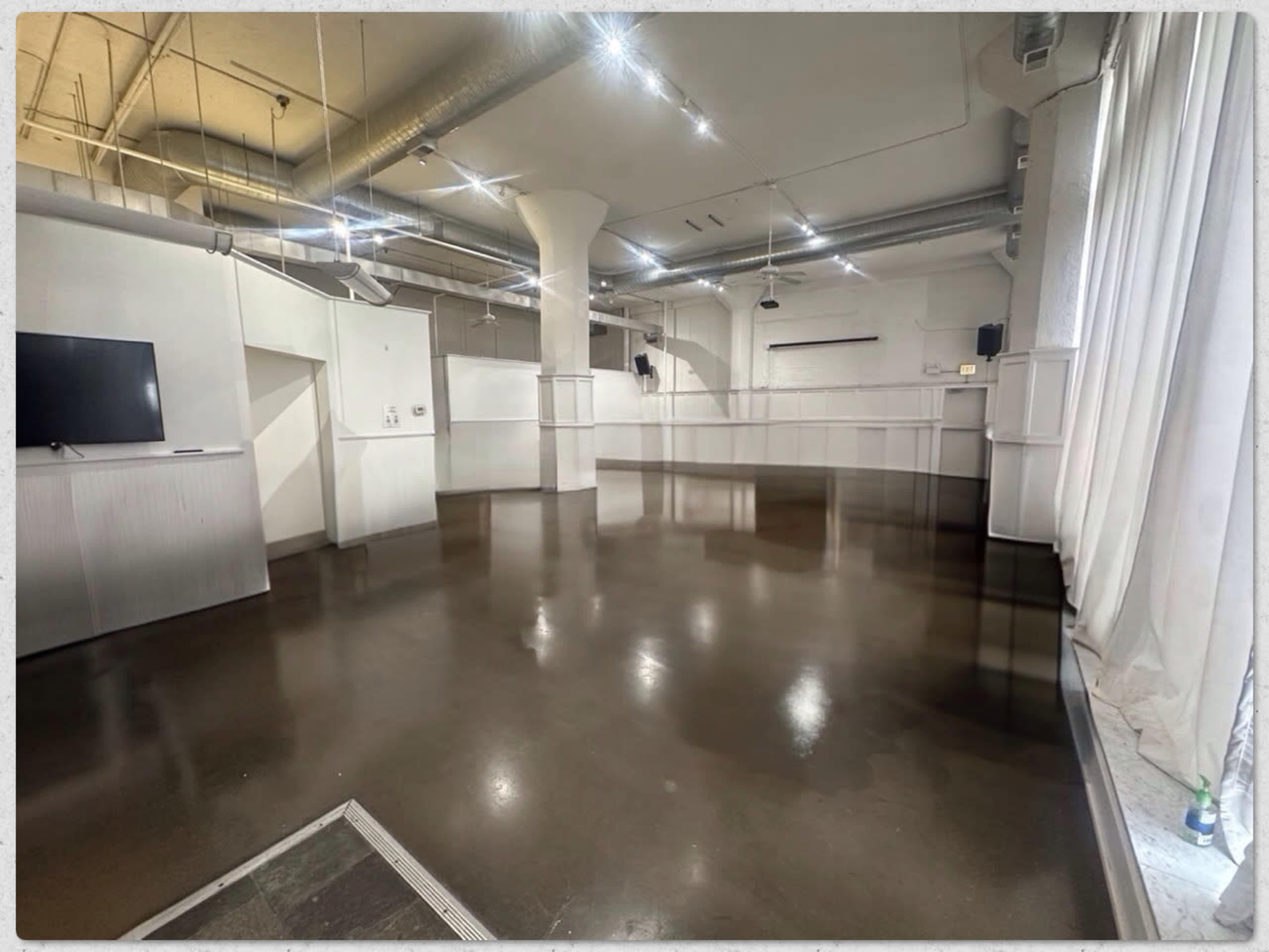 The image shows a large, empty room with polished concrete flooring, white walls, and several overhead lights.