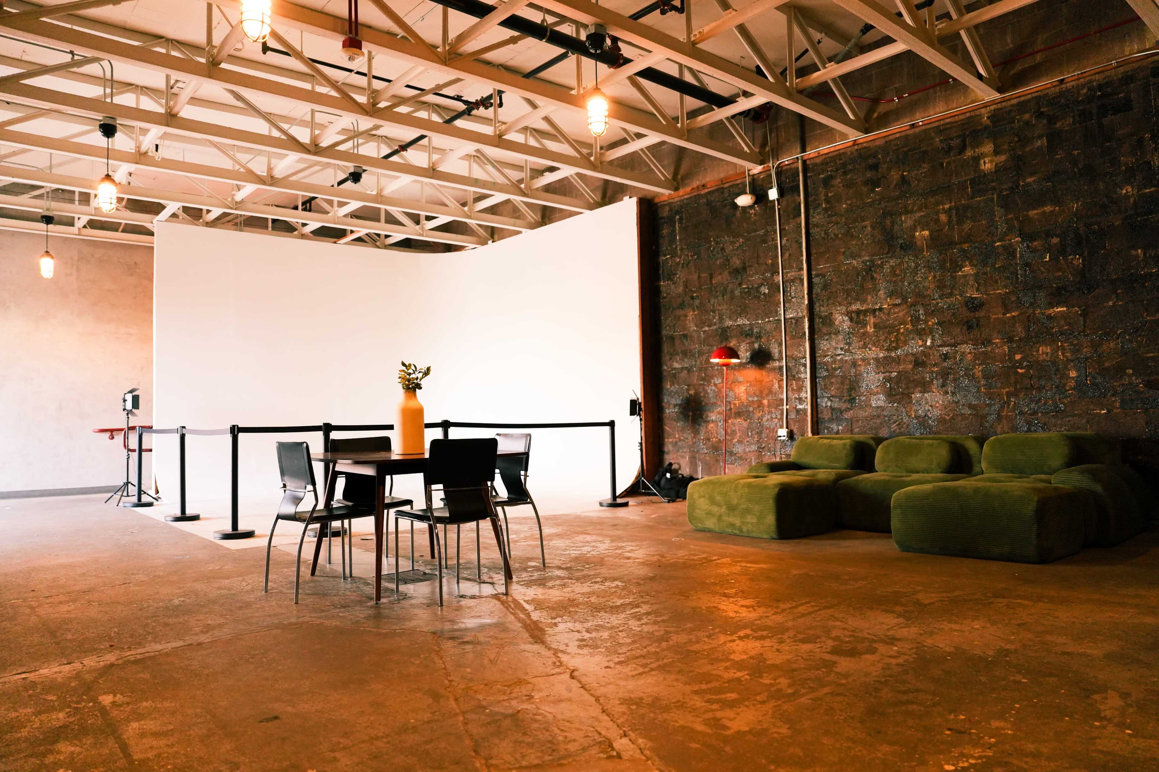 The image shows a spacious room with a large white backdrop, a wooden table and chairs in the center, and green seating along one wall.