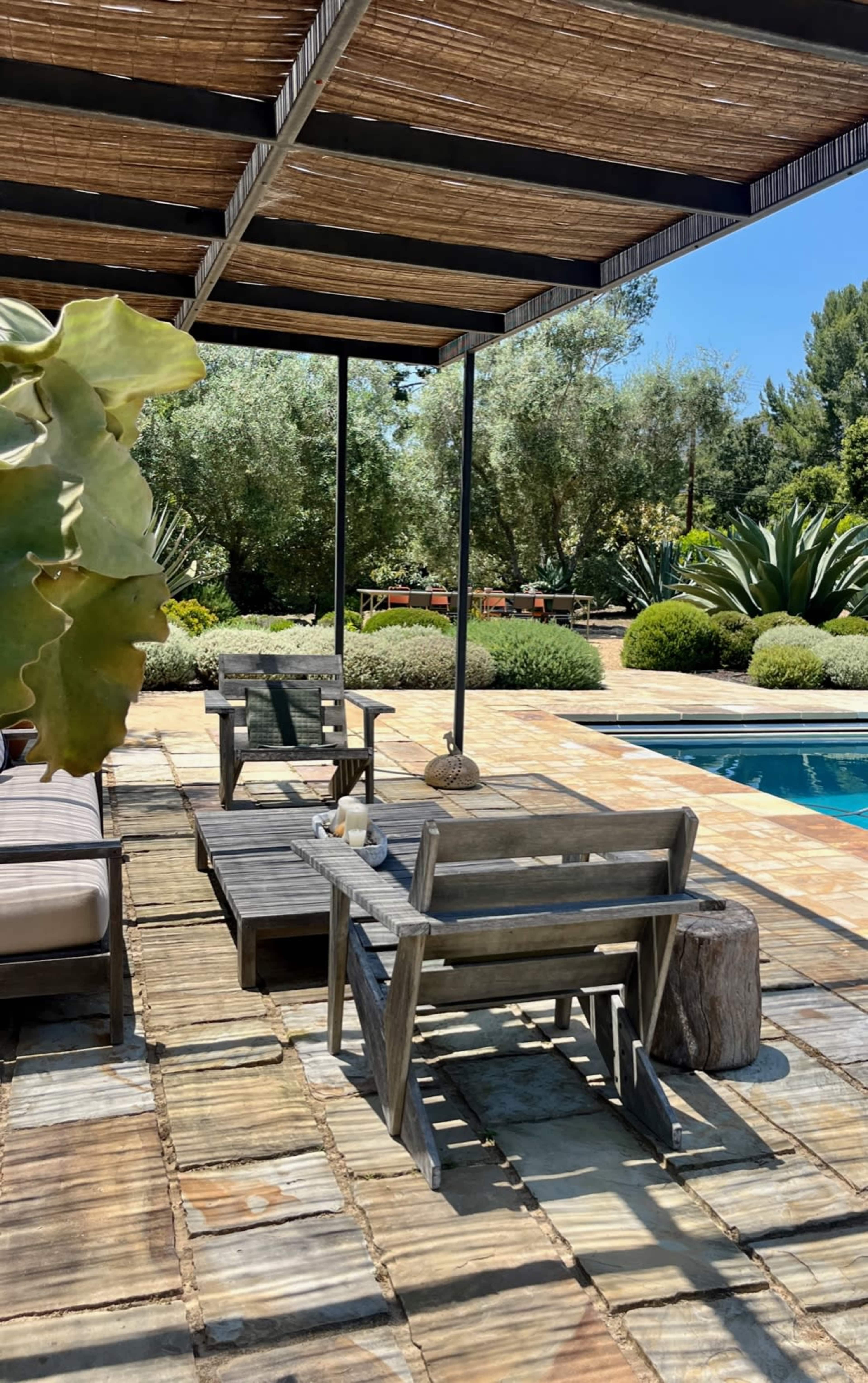 A wooden patio area features two chairs and a side table next to a swimming pool, surrounded by greenery and shaded by a roof of bamboo.