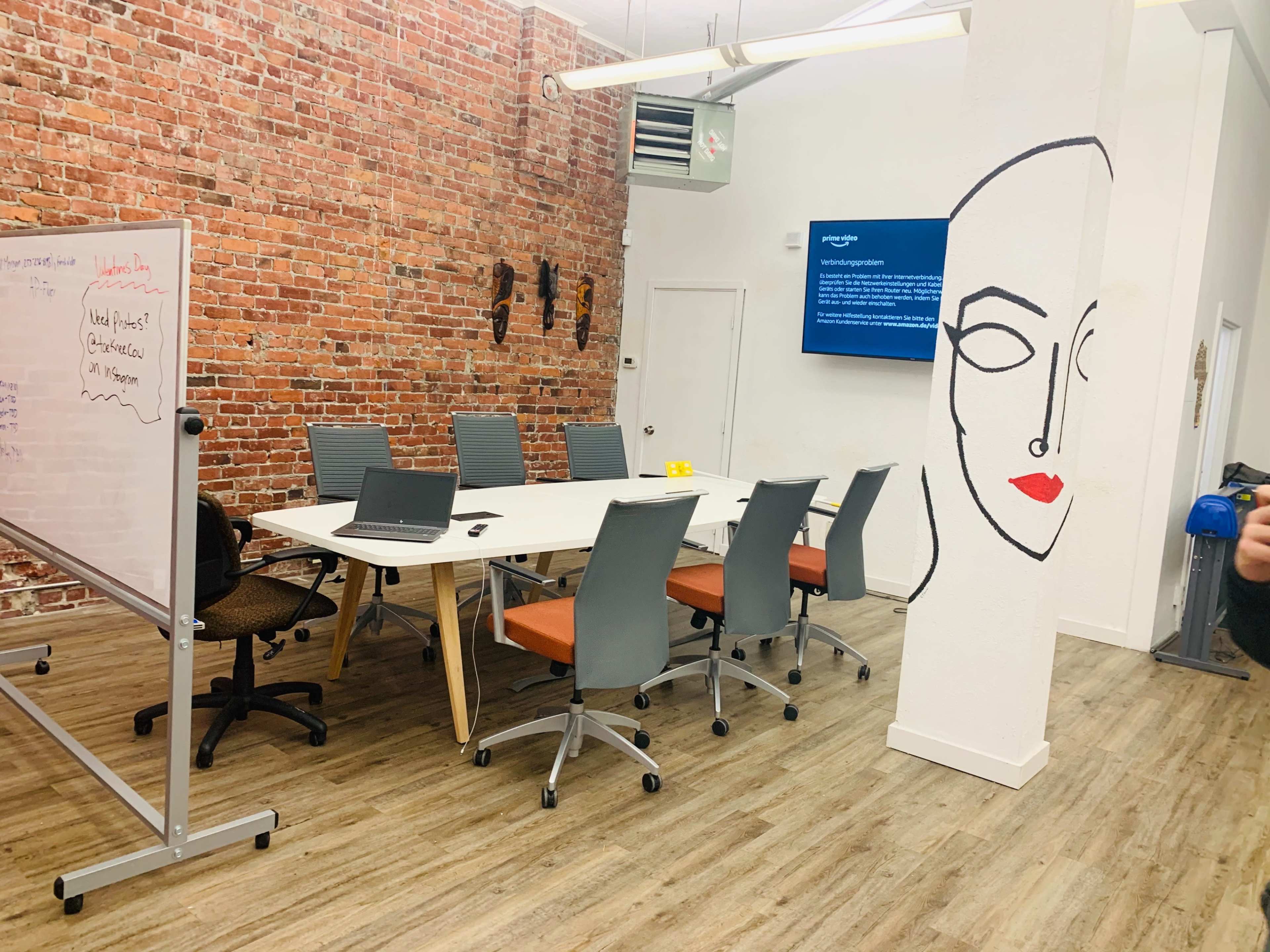 A modern conference room features a long table with several gray chairs, a whiteboard, and a wall adorned with a minimalist face drawing, set against a backdrop of exposed brick.