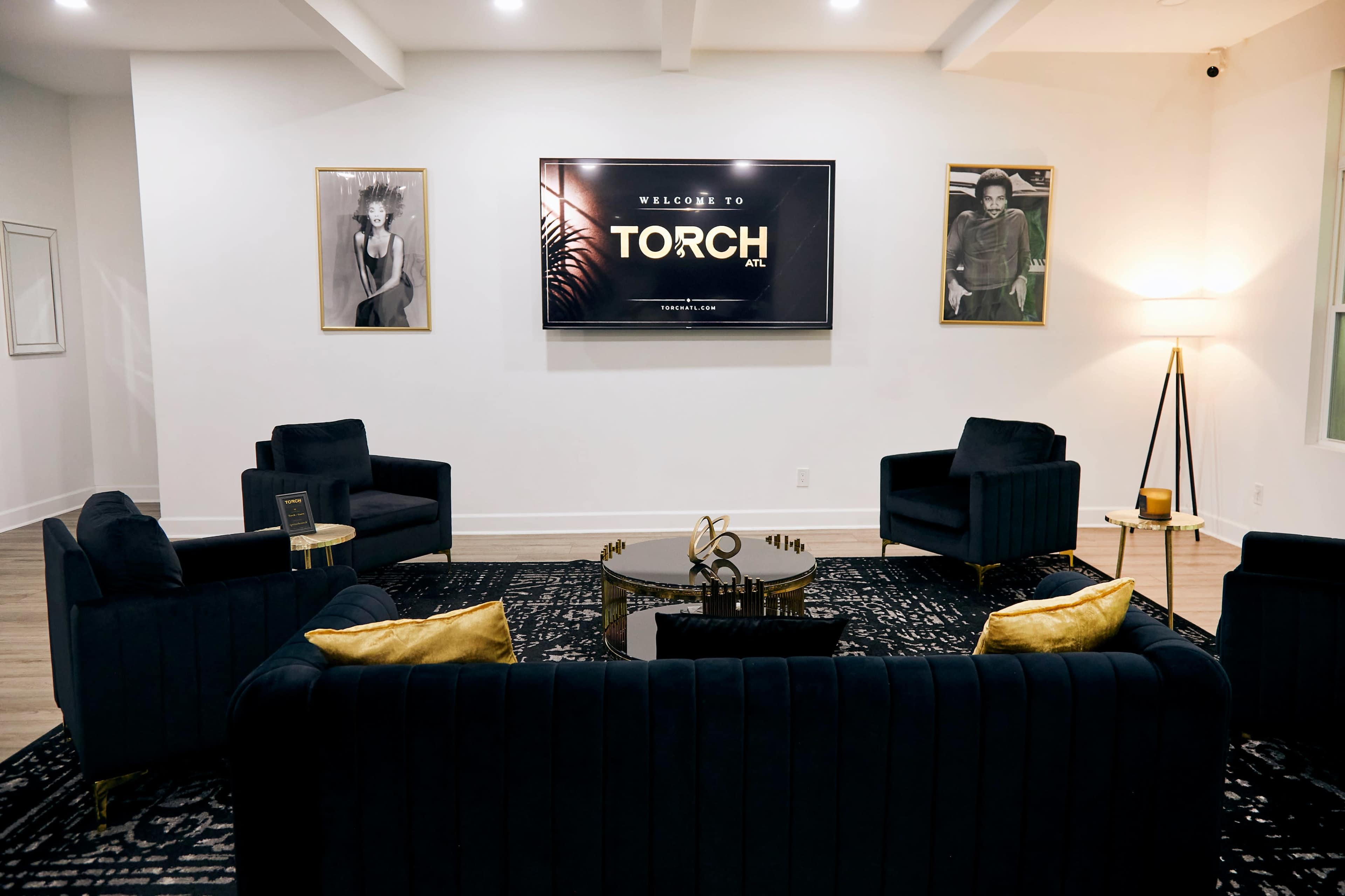 The image shows a modern lounge area featuring dark upholstered furniture arranged around a decorative coffee table, with a large screen displaying "WELCOME TO TORCH" on the wall and framed artwork hung on either side.