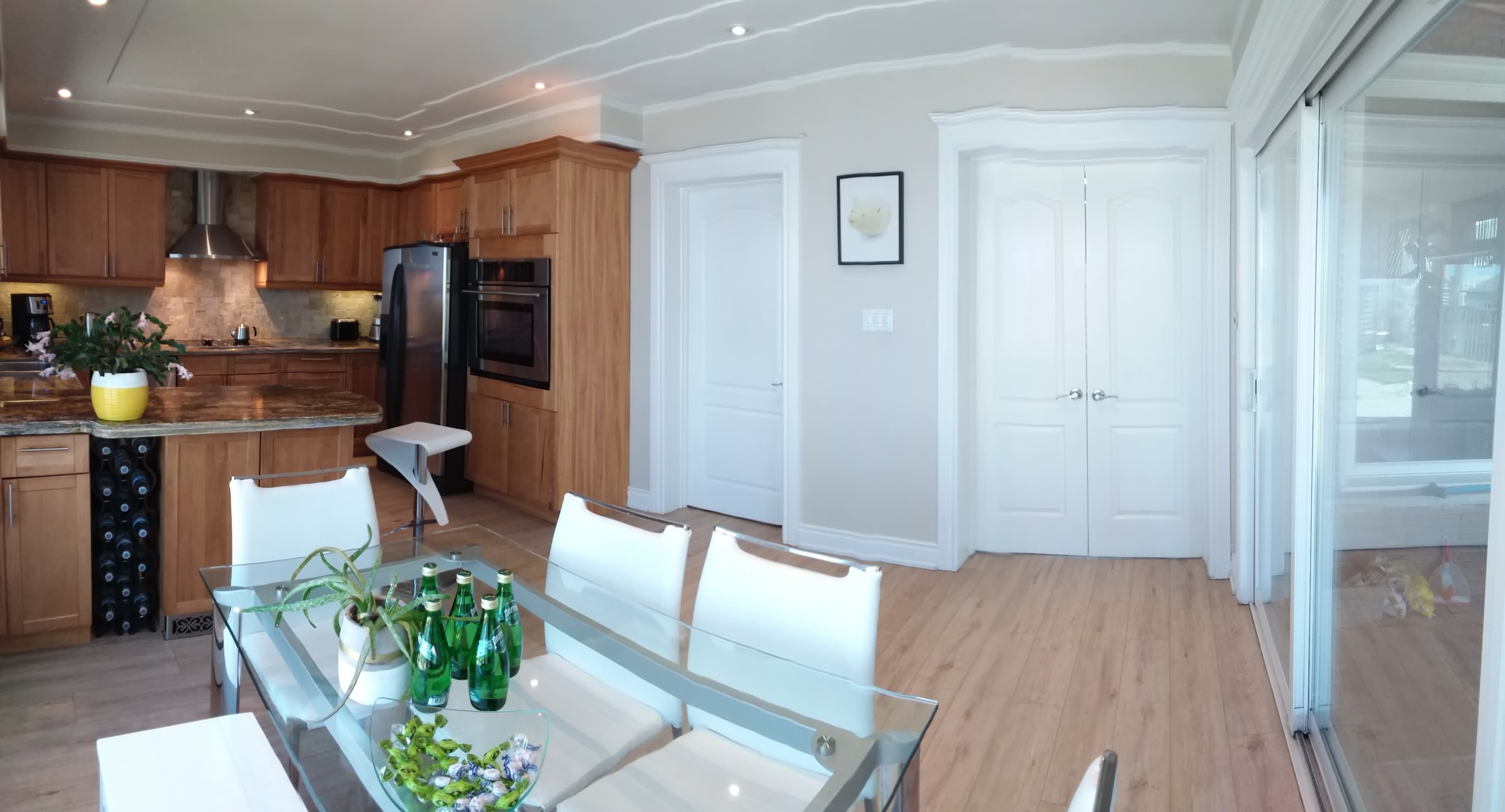 A spacious kitchen and dining area with wooden cabinets, a glass dining table set for four, and an adjoining room with large glass doors.