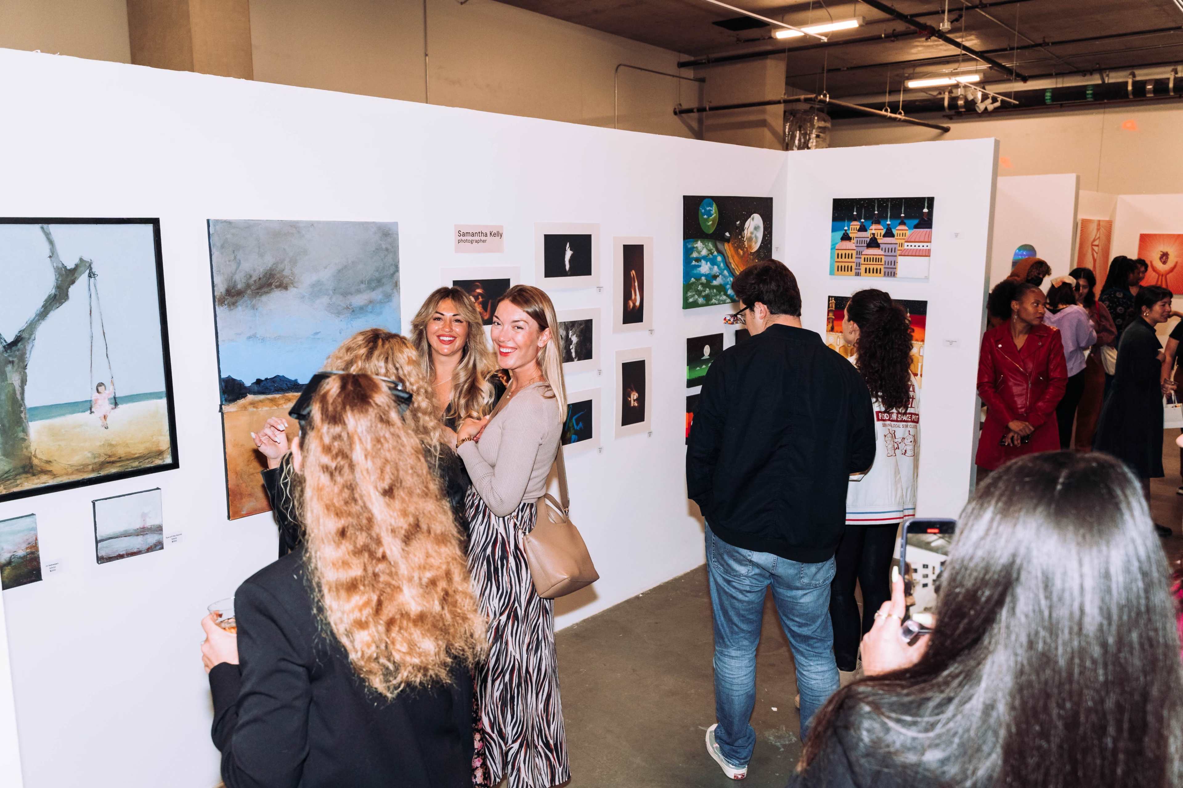 A group of people gather around an art exhibition showcasing various paintings on white walls.