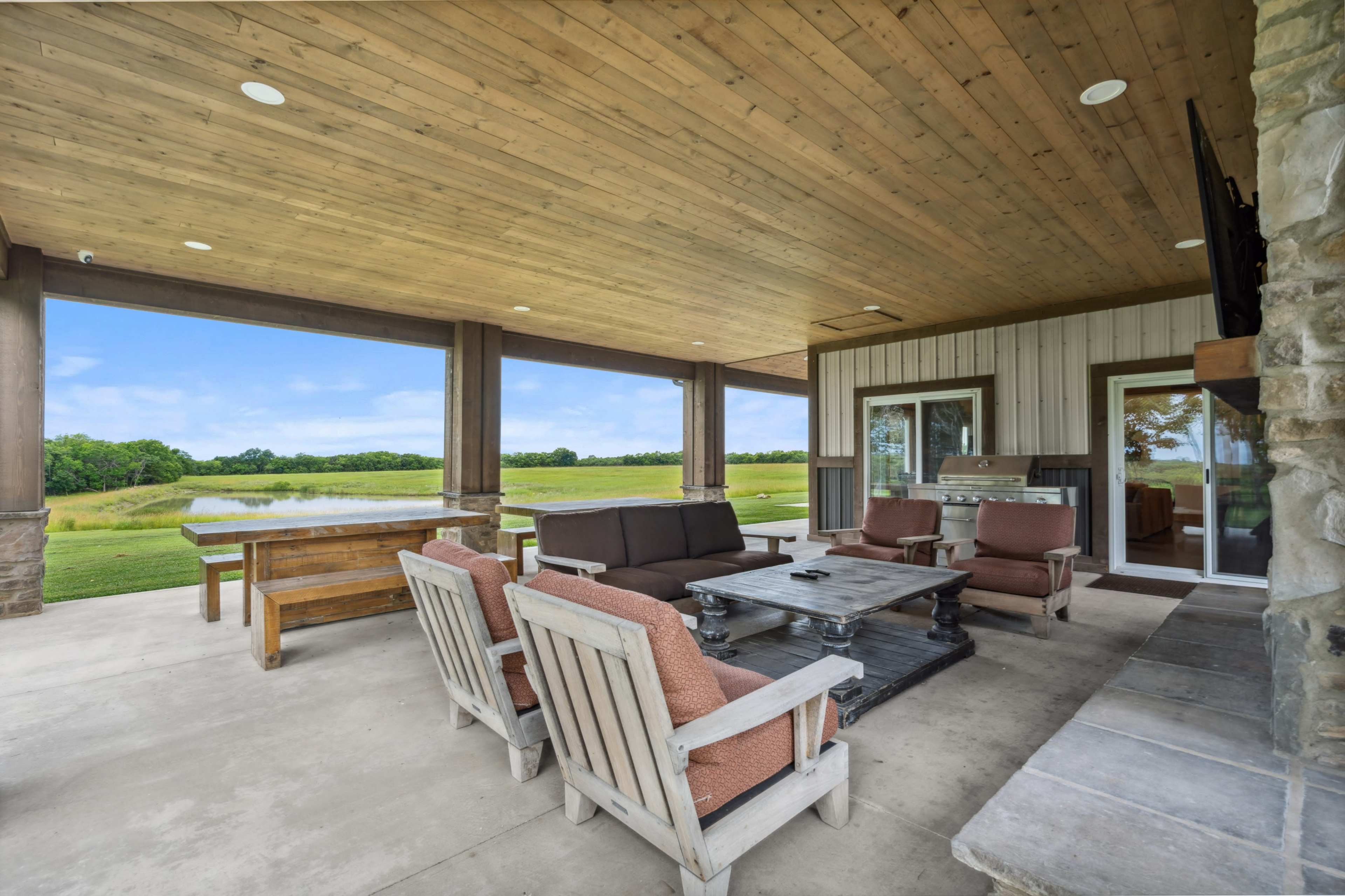 The image shows a covered outdoor seating area with wooden furniture, a stone fireplace, and a view of a grassy landscape.