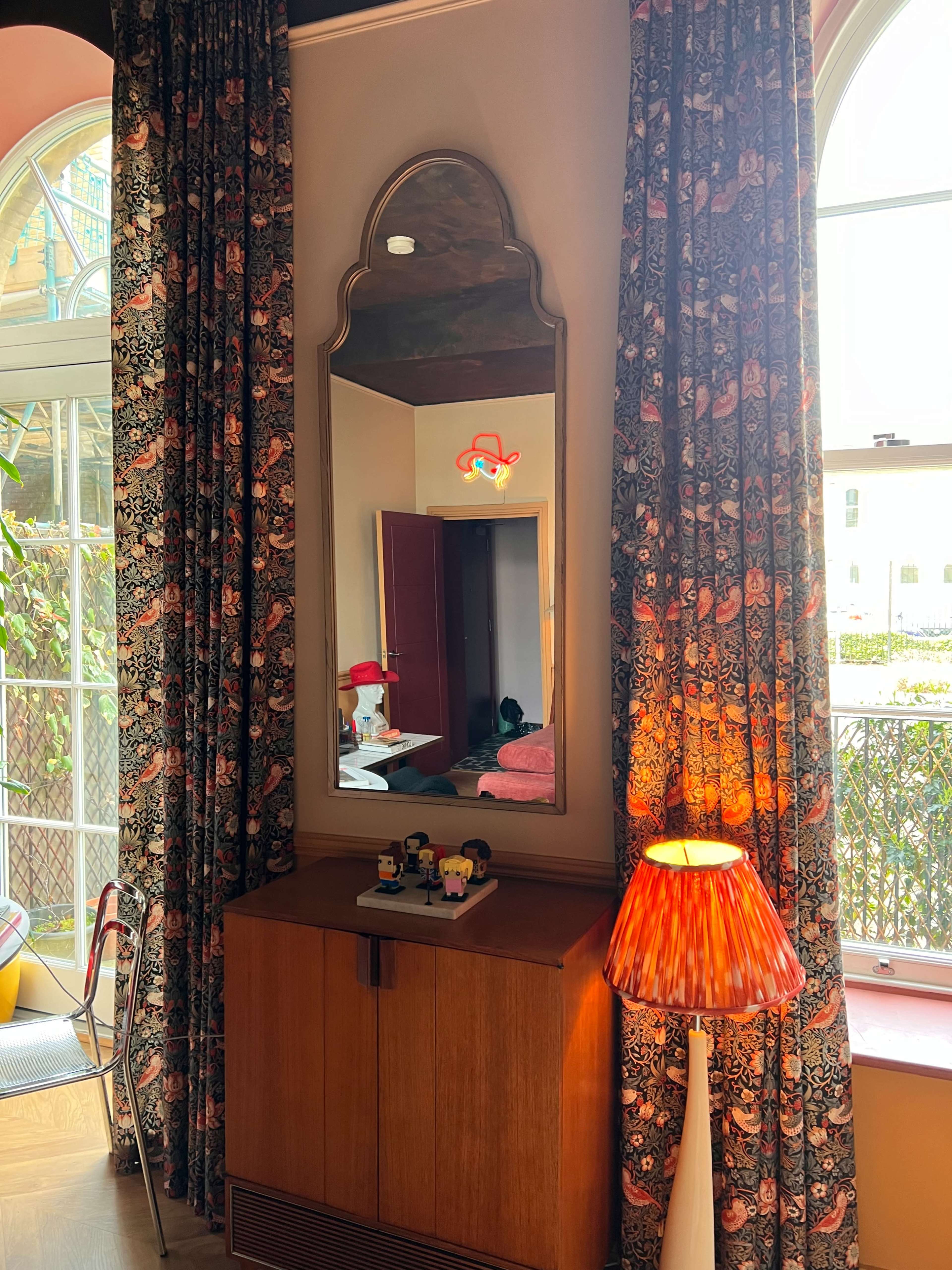 A room with a wooden cabinet under a decorative mirror, flanked by patterned curtains, with a lampshade casting light and a view of greenery outside.