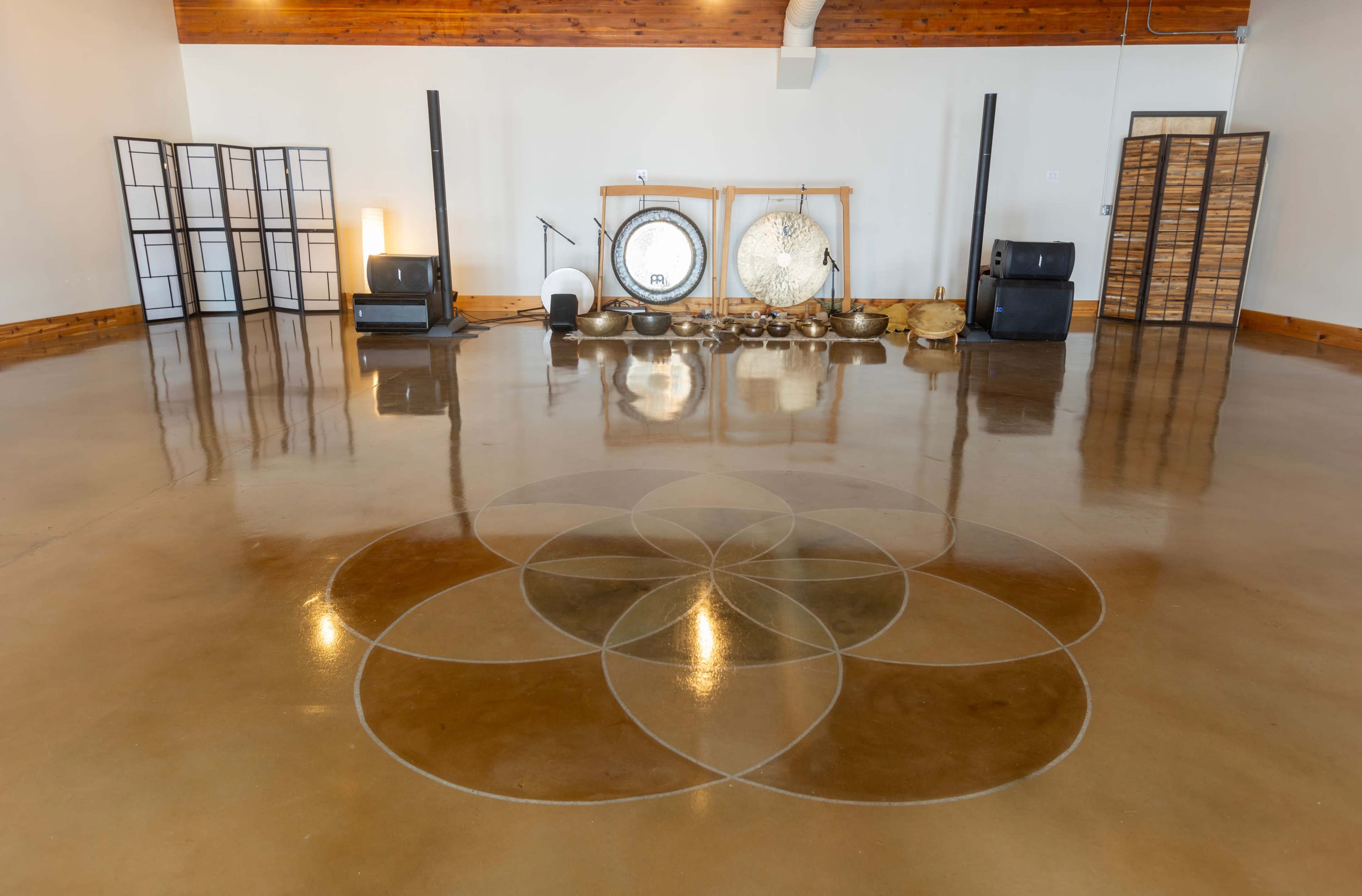 A spacious meditation or wellness studio with polished concrete flooring featuring a circular design, surrounded by various sound healing instruments along the back wall.