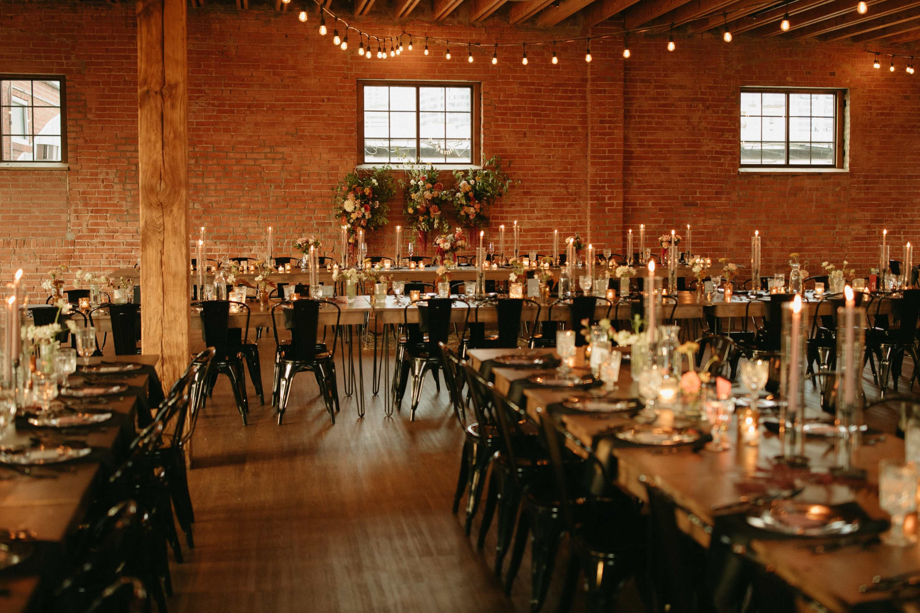 A spacious event hall with wooden beams and brick walls, featuring elegantly set dining tables adorned with floral arrangements and candles, and illuminated by string lights overhead.