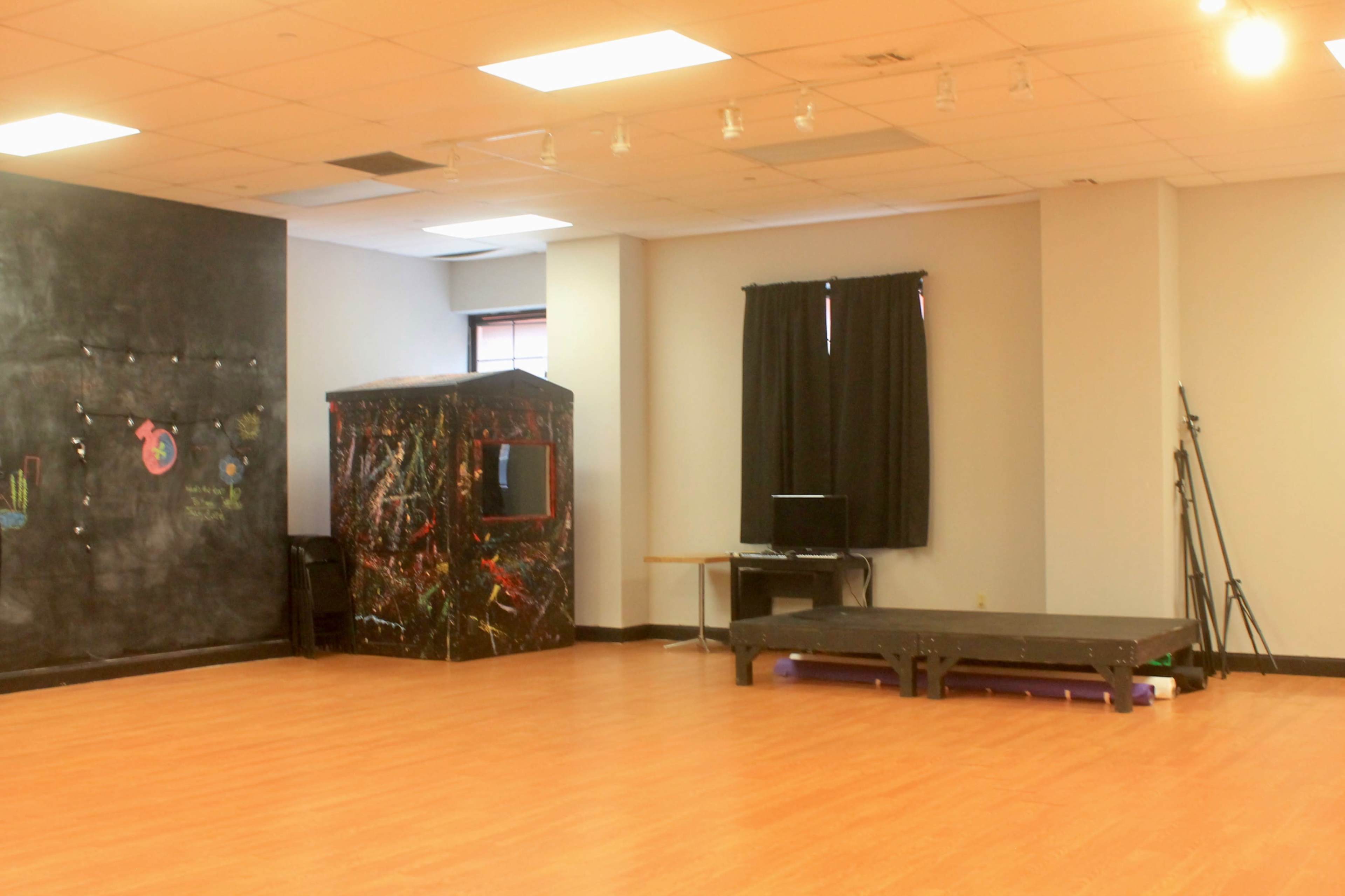 A spacious studio with wooden flooring, featuring a performance stage, a black wall with colorful artwork, and a dark curtain on one side.