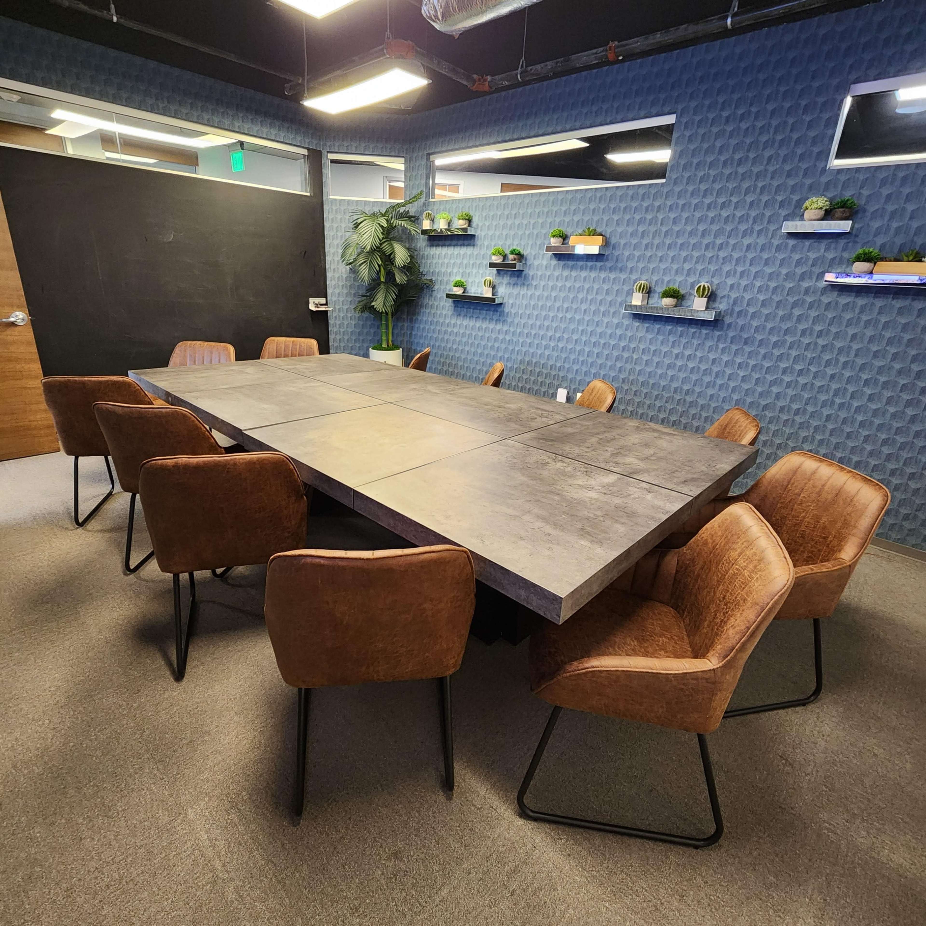 A large, rectangular conference table surrounded by eight brown chairs is positioned in a modern meeting room with blue textured walls and small potted plants on shelves.