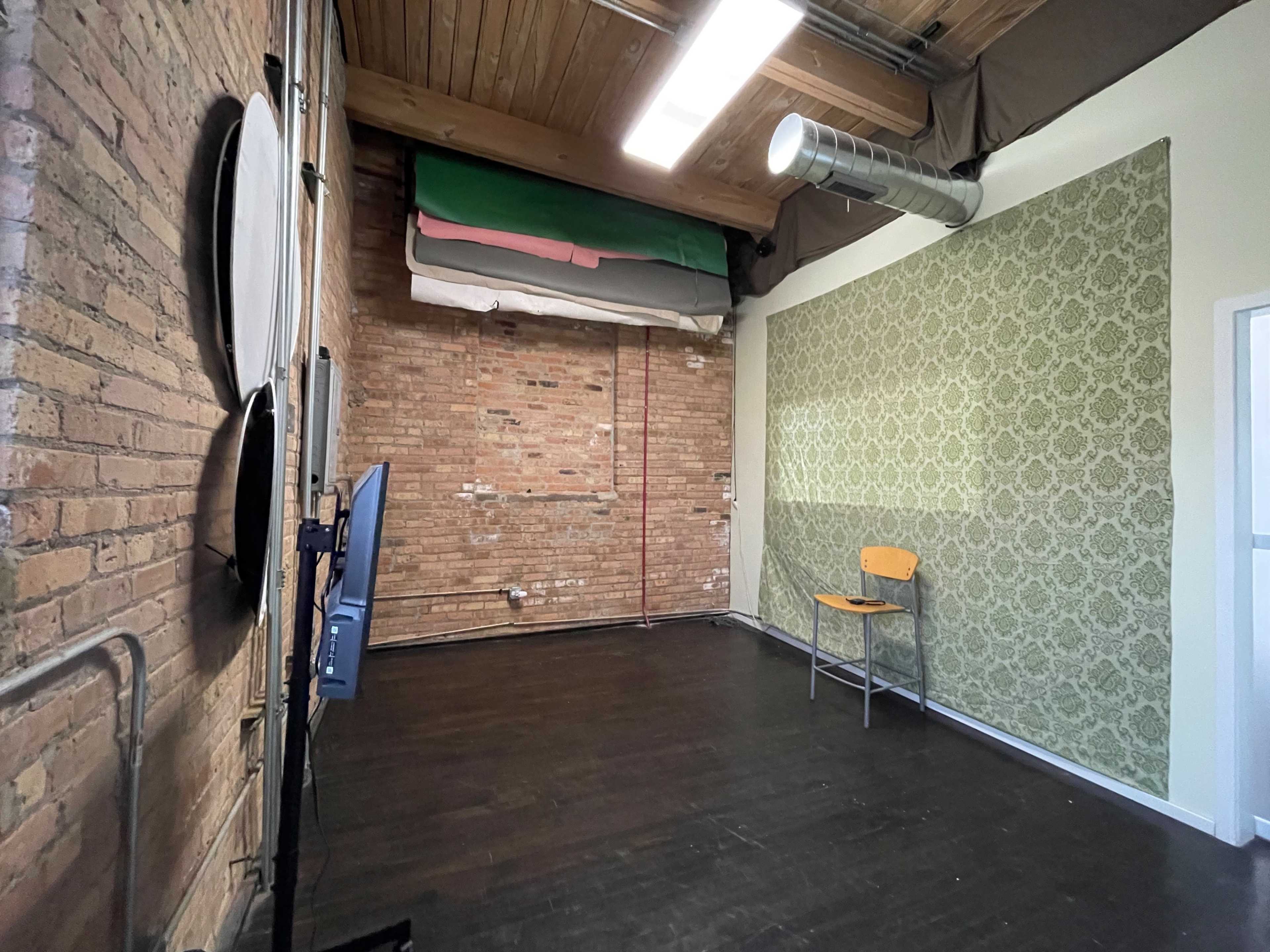 The image shows a room with exposed brick walls, a patterned green accent wall, a wooden chair, and a television screen on a stand.