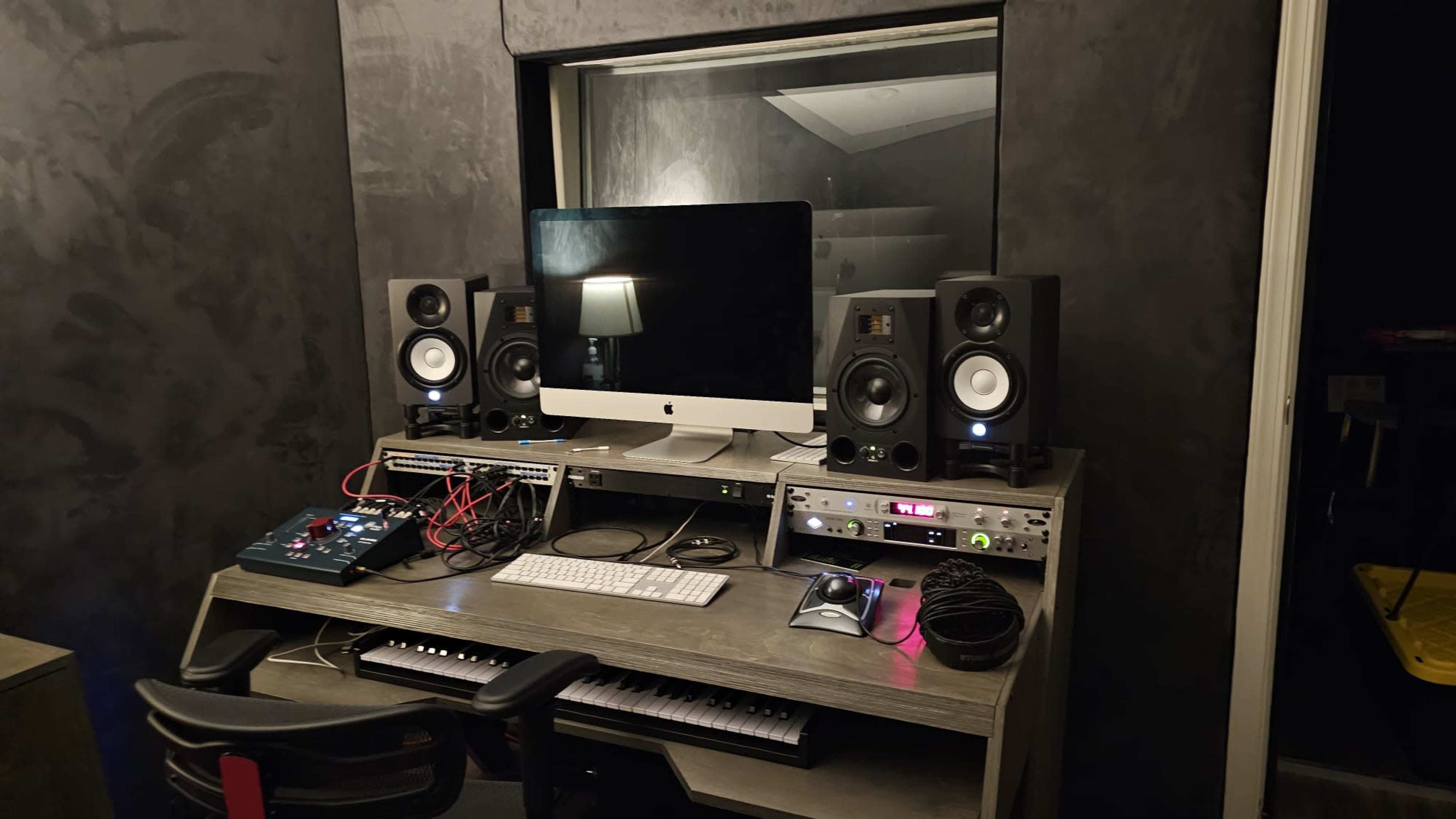 A music production studio features an iMac, studio monitors, audio interfaces, and a keyboard arranged on a desk.
