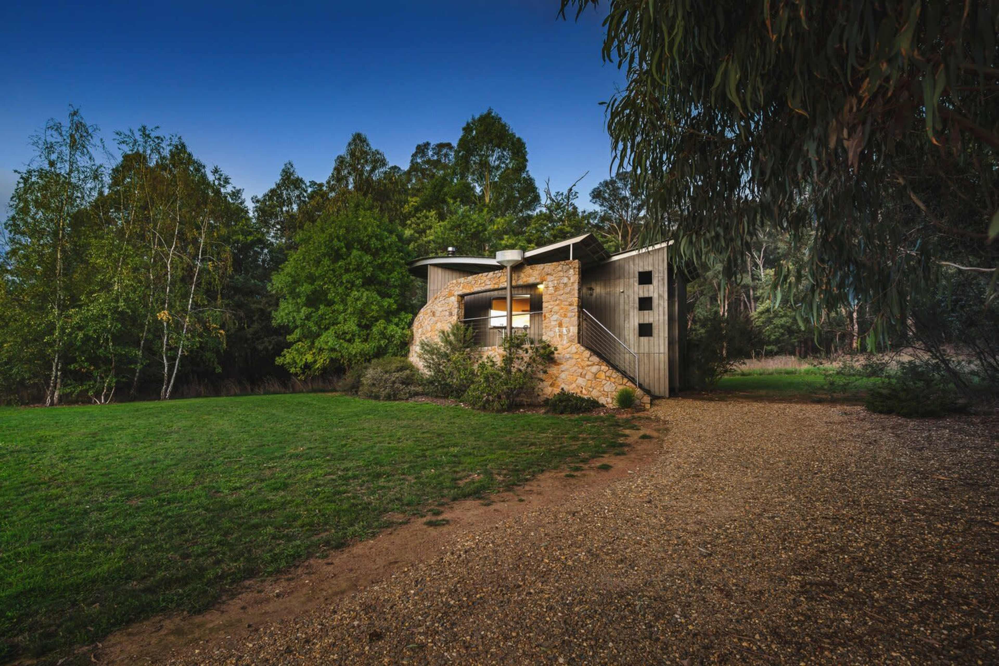 A modern house with a stone facade set among trees on a grassy landscape, with a gravel path leading to its entrance.