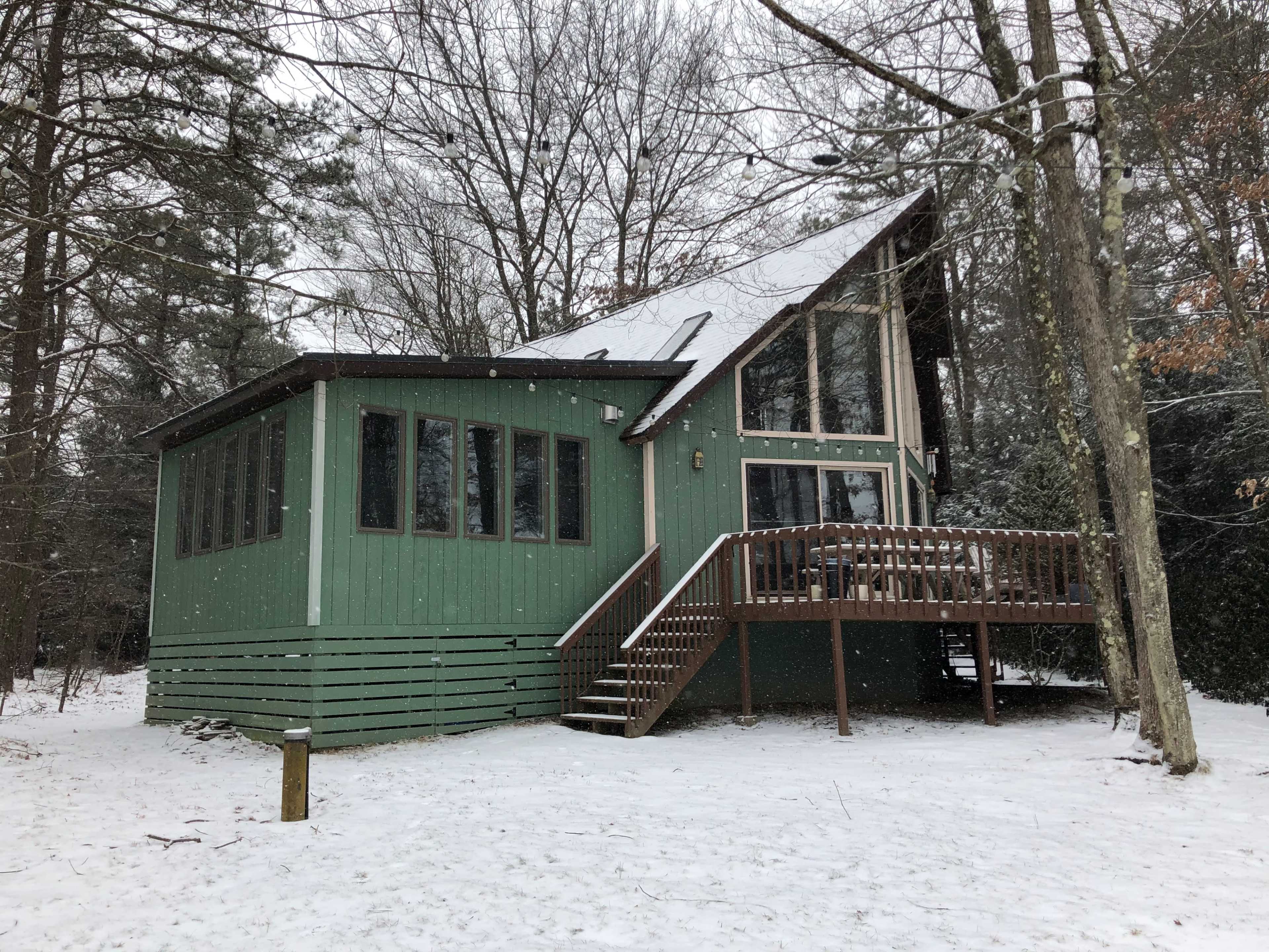 A green wooden cabin with a sloped roof sits in a snow-covered forest clearing.