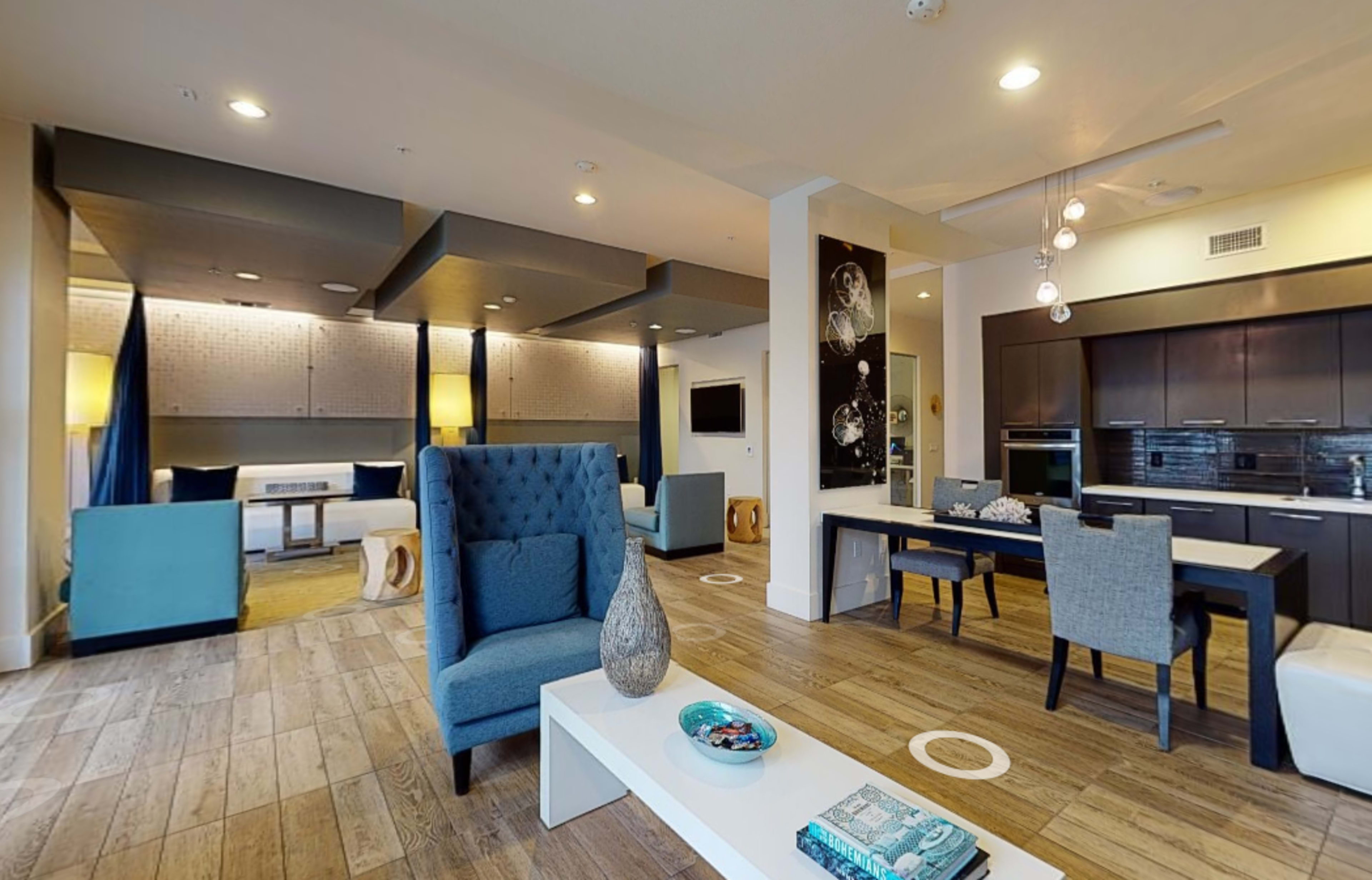 A modern living space featuring a blue armchair, a dining area with gray chairs, and a kitchen in the background, all with wooden flooring.