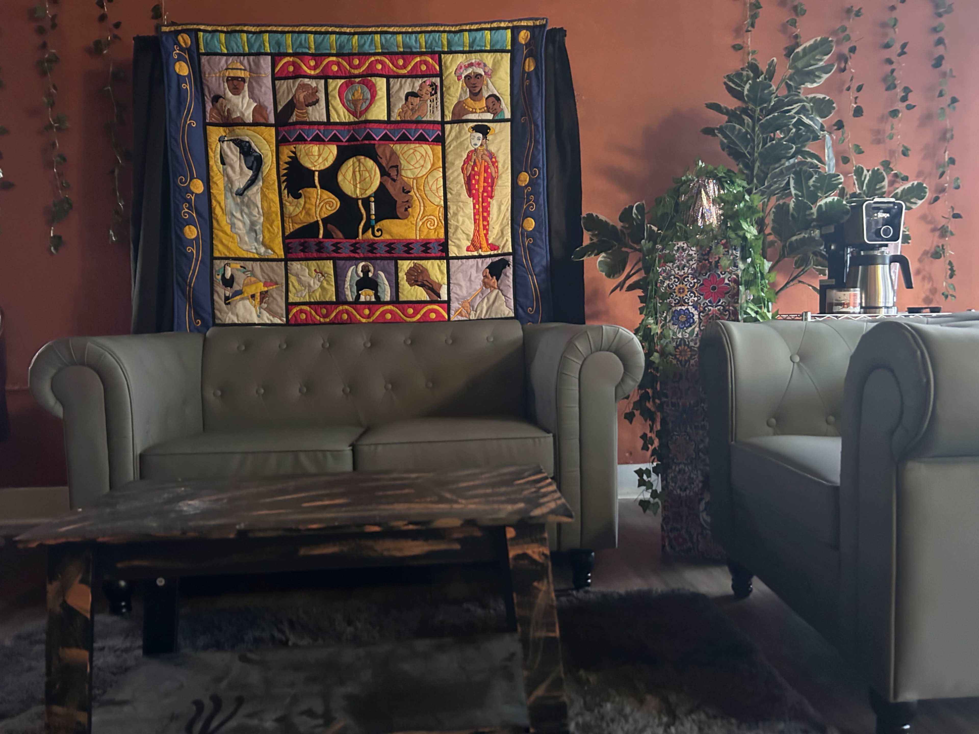 The scene features a cozy seating area with two tufted sofas, a patterned wall hanging, and a coffee table, surrounded by plants and decor against a warm-colored wall.