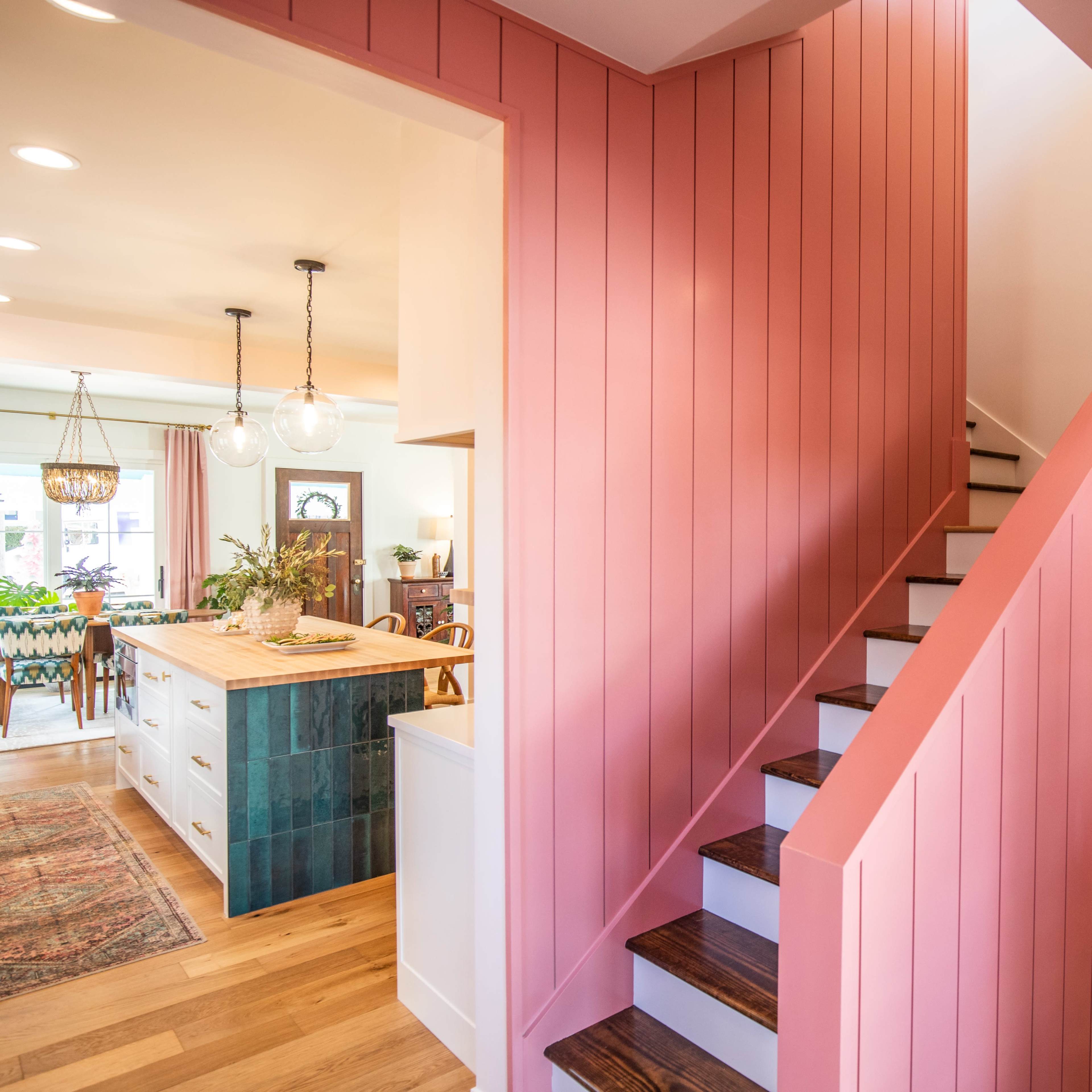 A bright hallway with pink-paneled walls leads to a staircase, with a view of a kitchen featuring a wooden island and teal-tiled accents.